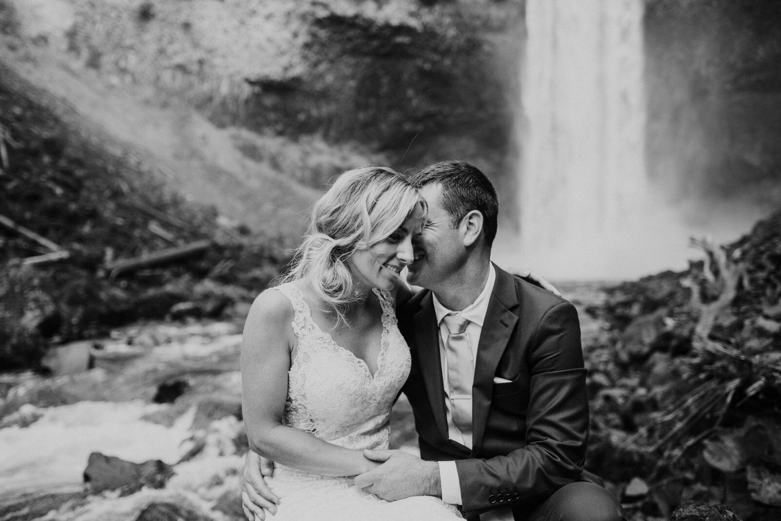 groom whispering into brides ear sitting below waterfall