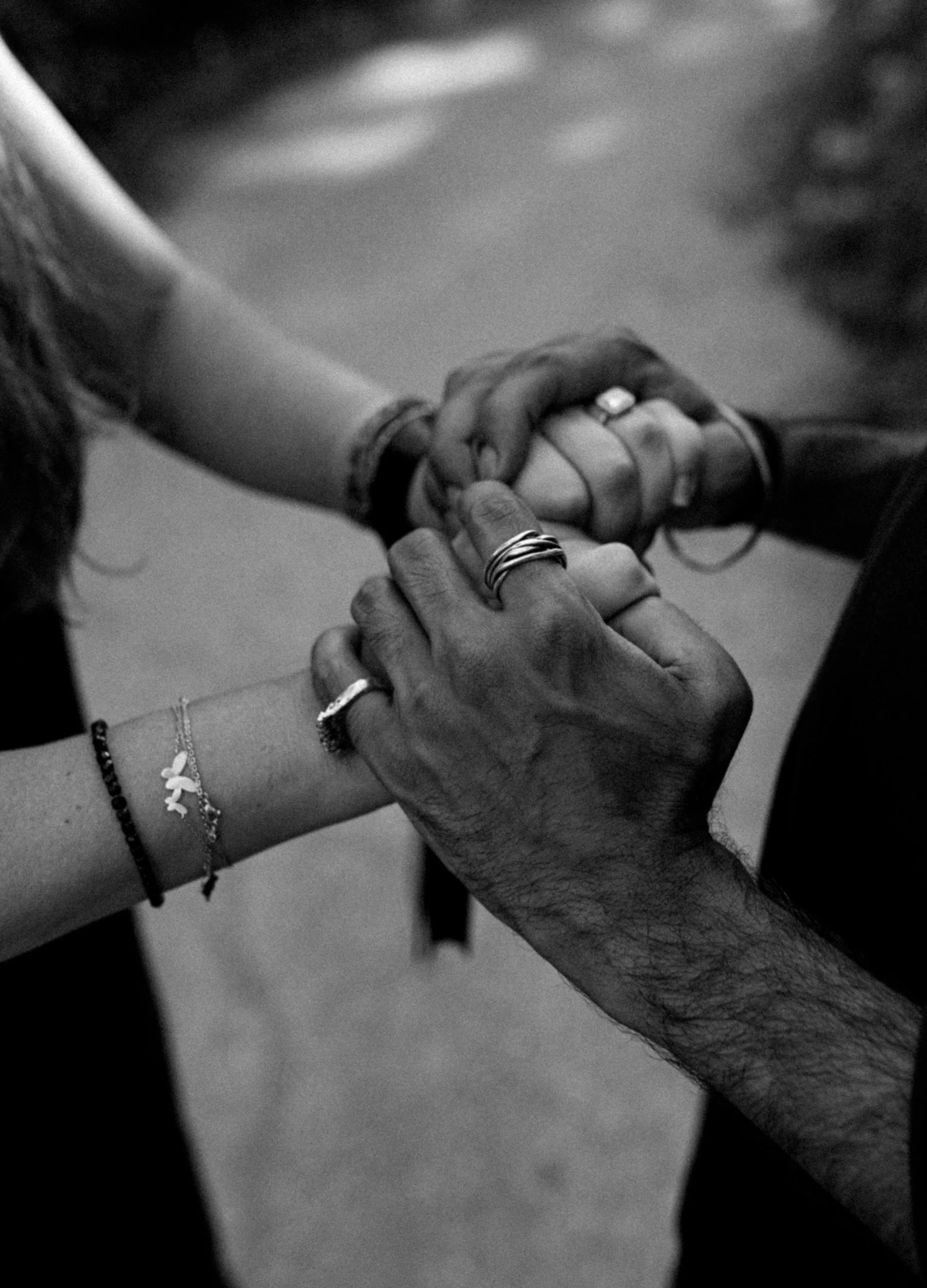 A black and white photo of four hands intertwined and holding each other, two are male hands and two are female hands, with rings and bracelets visible.