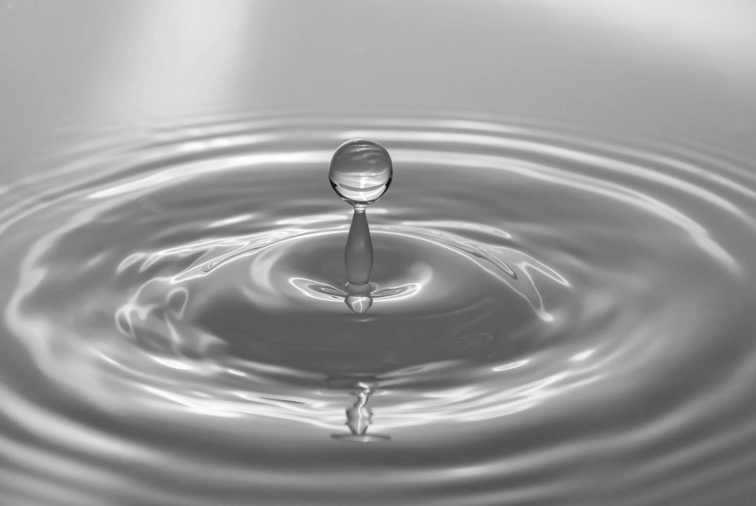 Black and white photo of a water droplet splashing into a calm water surface, creating ripples and a vertical splash.