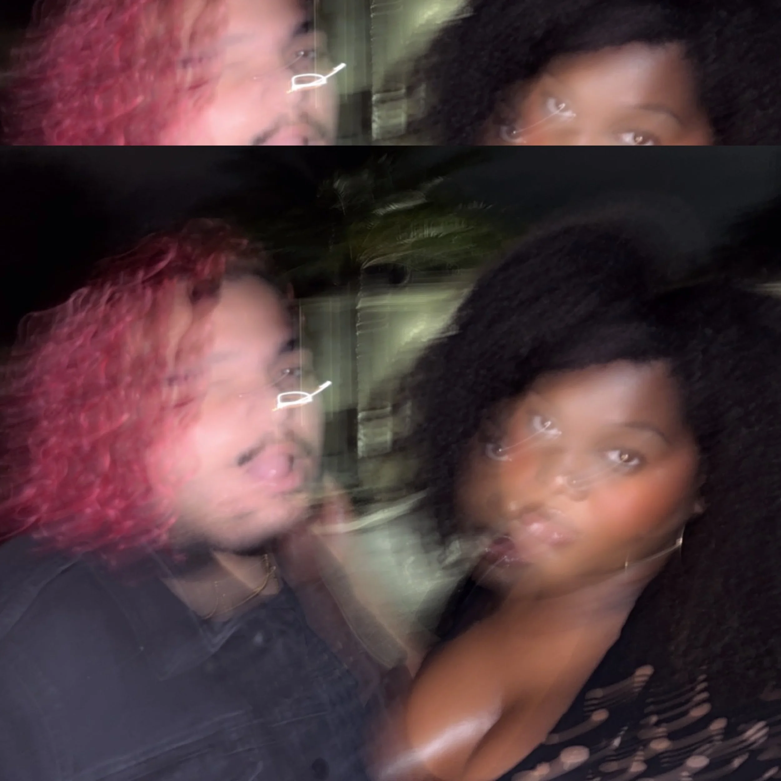 Two people with curly hair, one with pink hair, and the other with black hair, are close together in a blurry, low-light photo.