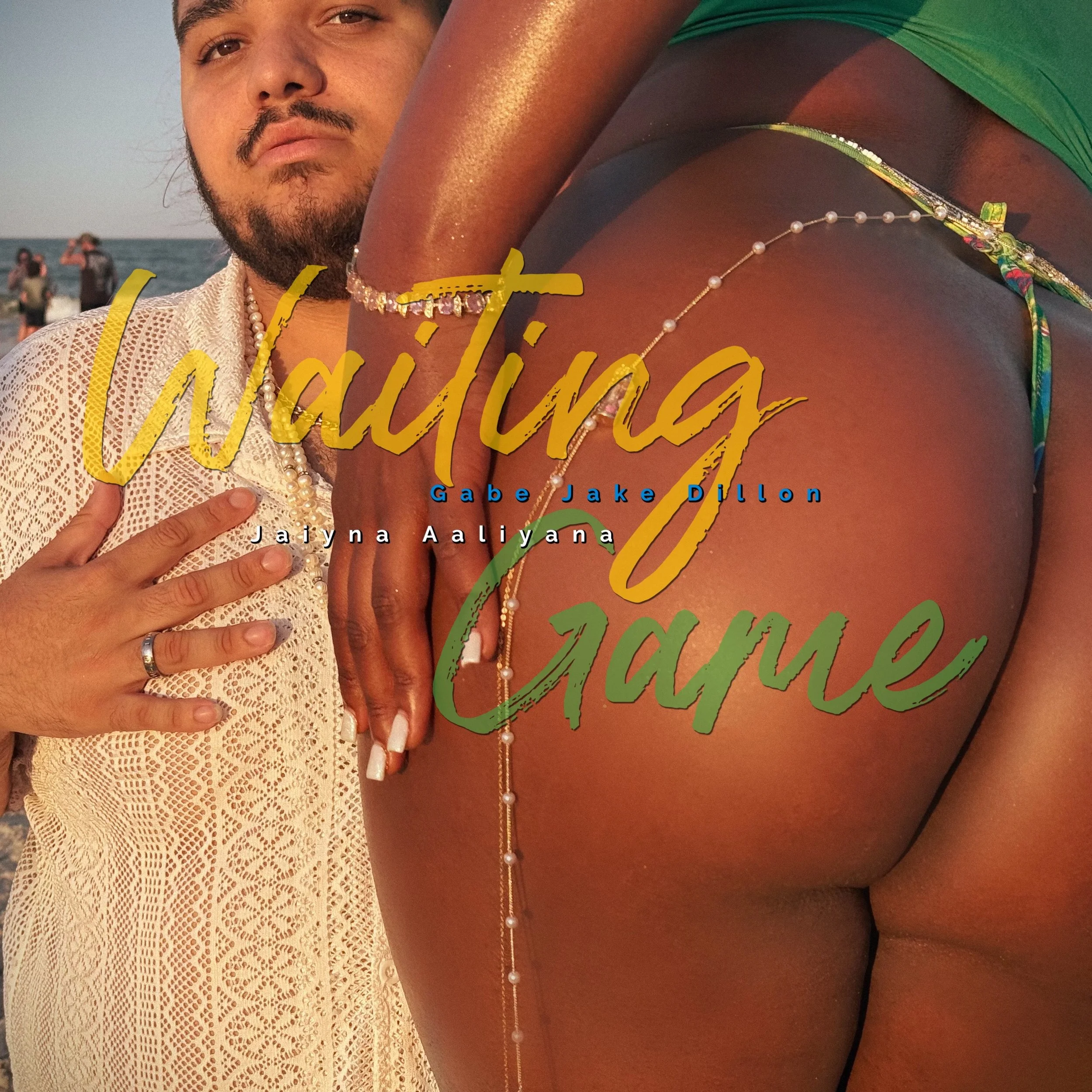 A man stands on a beach with an arm around a woman in a green bikini, embracing her from behind. The man's hand rests on his chest, and he is wearing a white lace shirt. The woman is wearing jewelry, including a pearl belly chain. The beach scene is in the background with other people visible near the water.