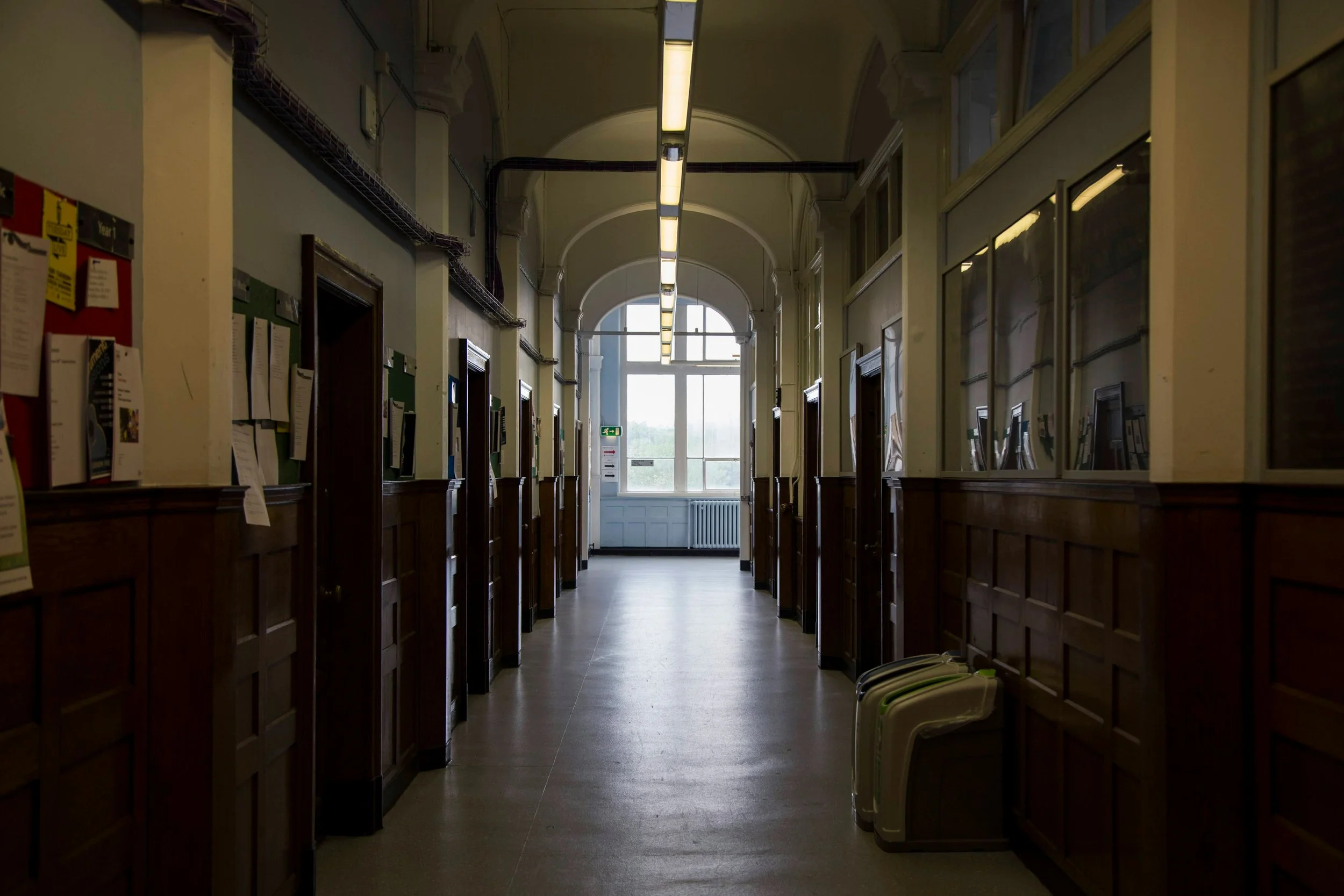 Empty hallway in a building with wood paneling, bulletin boards on the walls, and large windows at the end.
