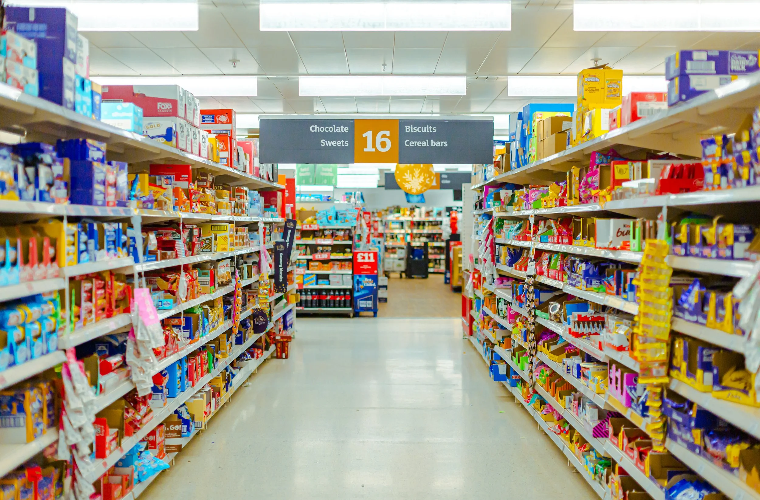 Aisle in a grocery store with shelves stocked with candy, cookies, and snack items, and a sign overhead indicating aisle 16 for chocolate, sweets, biscuits, and cereal bars.
