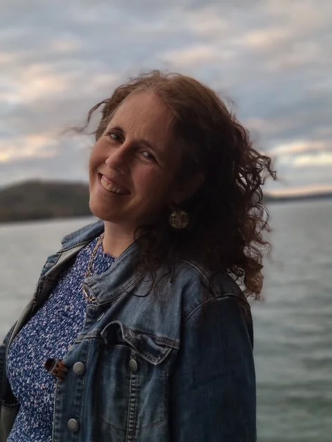 Woman with glasses and curly hair taking a selfie by a lake during sunset, with a dock extending into the water in the background.