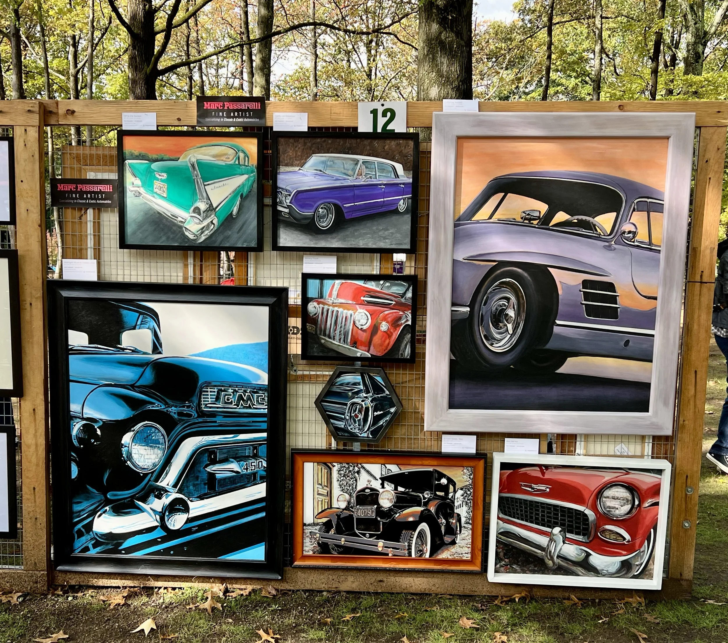 Display of colorful car paintings at an outdoor art fair, with a wood fence and trees in the background.