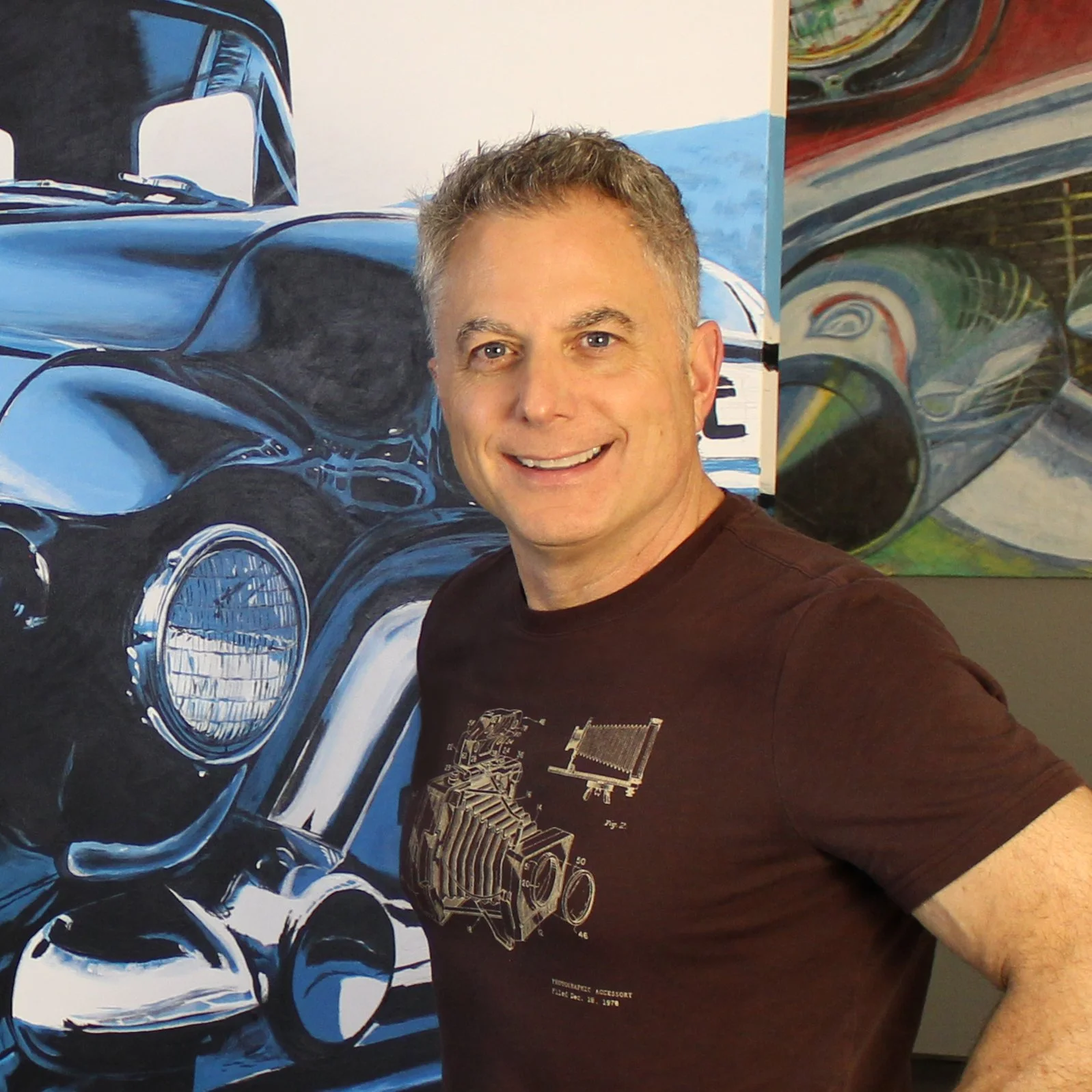 A smiling man with short hair taking a selfie in front of a large painting of a blue vintage car. The man is wearing a dark t-shirt with a technical diagram.