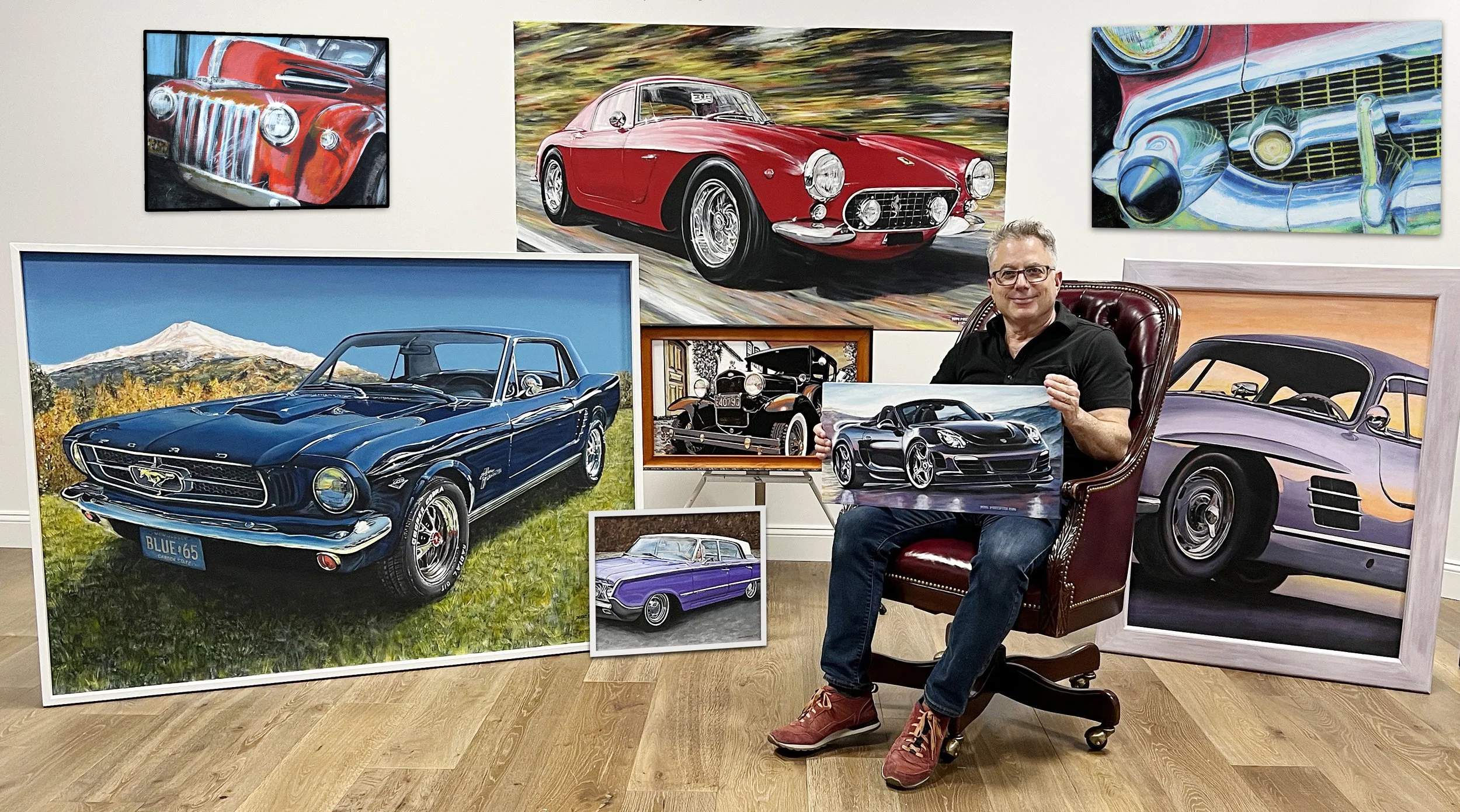 A man sitting in a leather armchair in a room decorated with numerous paintings of classic cars, including a red vintage sports car, a blue muscle car, a purple coupe, and a black vintage automobile, all displayed on the wall and floor.