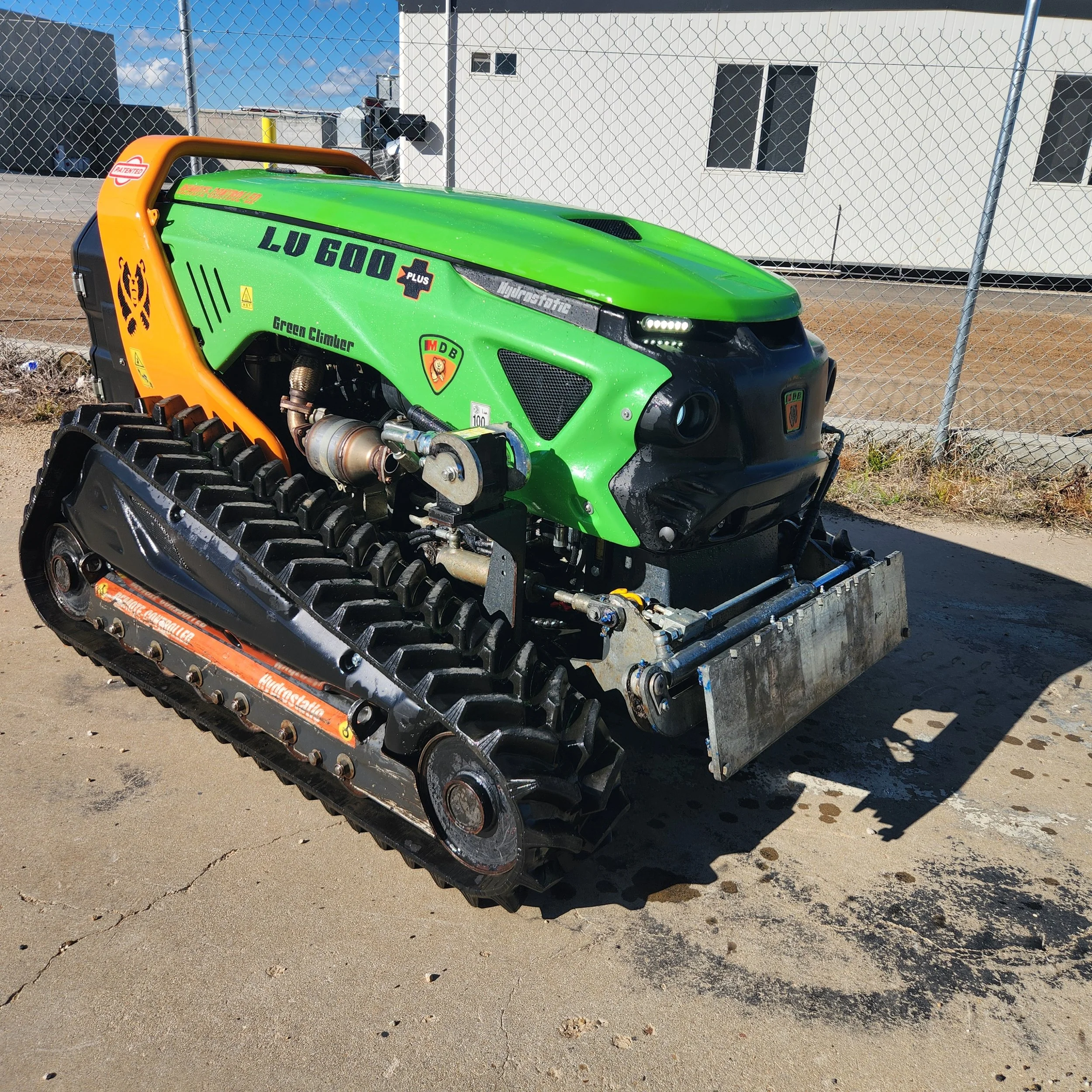 A green tracked off-road vehicle with black tracks, labeled 'LUV 600 Plus,' parked outdoors on a concrete surface. It has various stickers and a front attachment, with a chain-link fence and a building in the background.