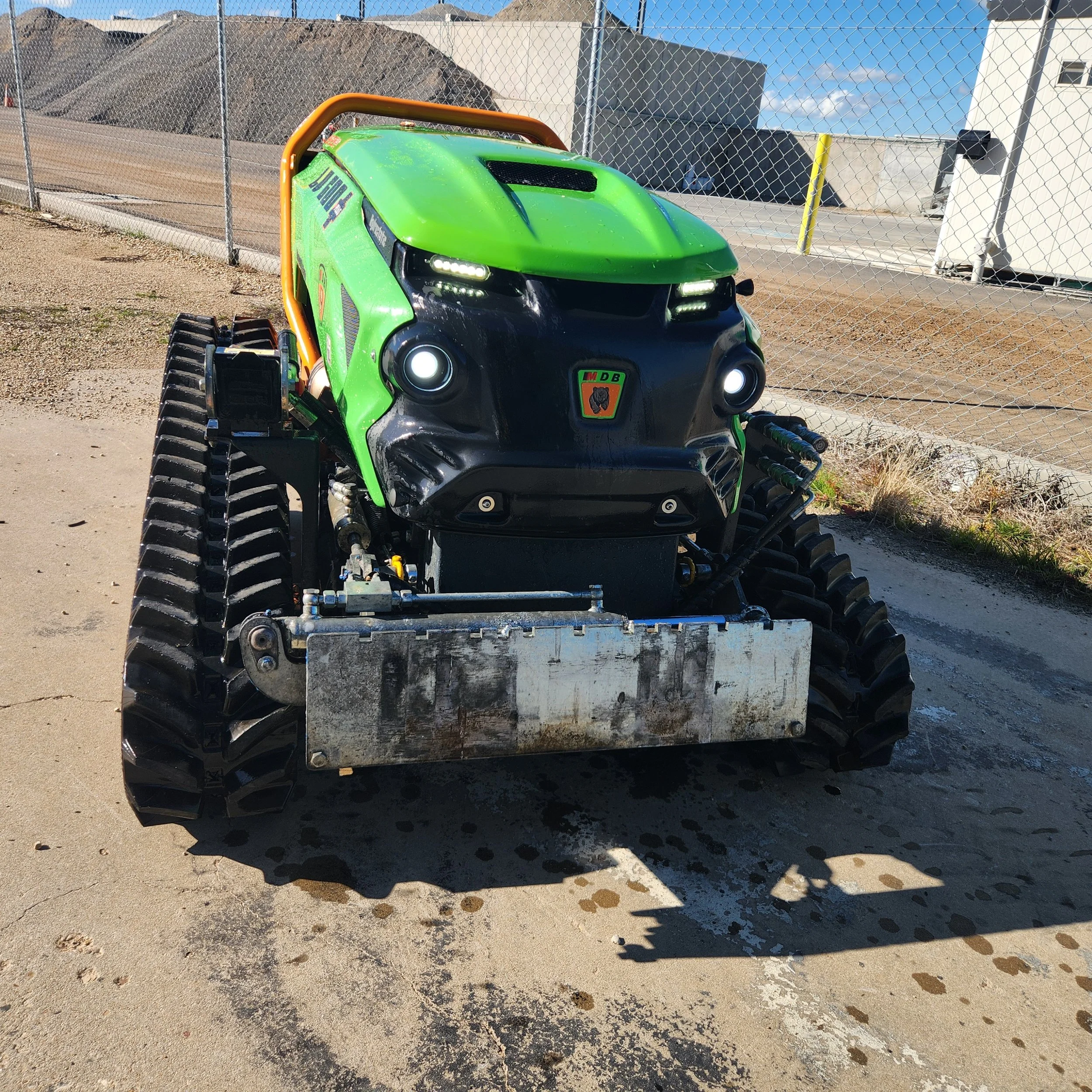 Autonomous robotic snow plow with green and black body, tracked wheels, and a metal blade in front, parked on a dirt surface near a chain-link fence.