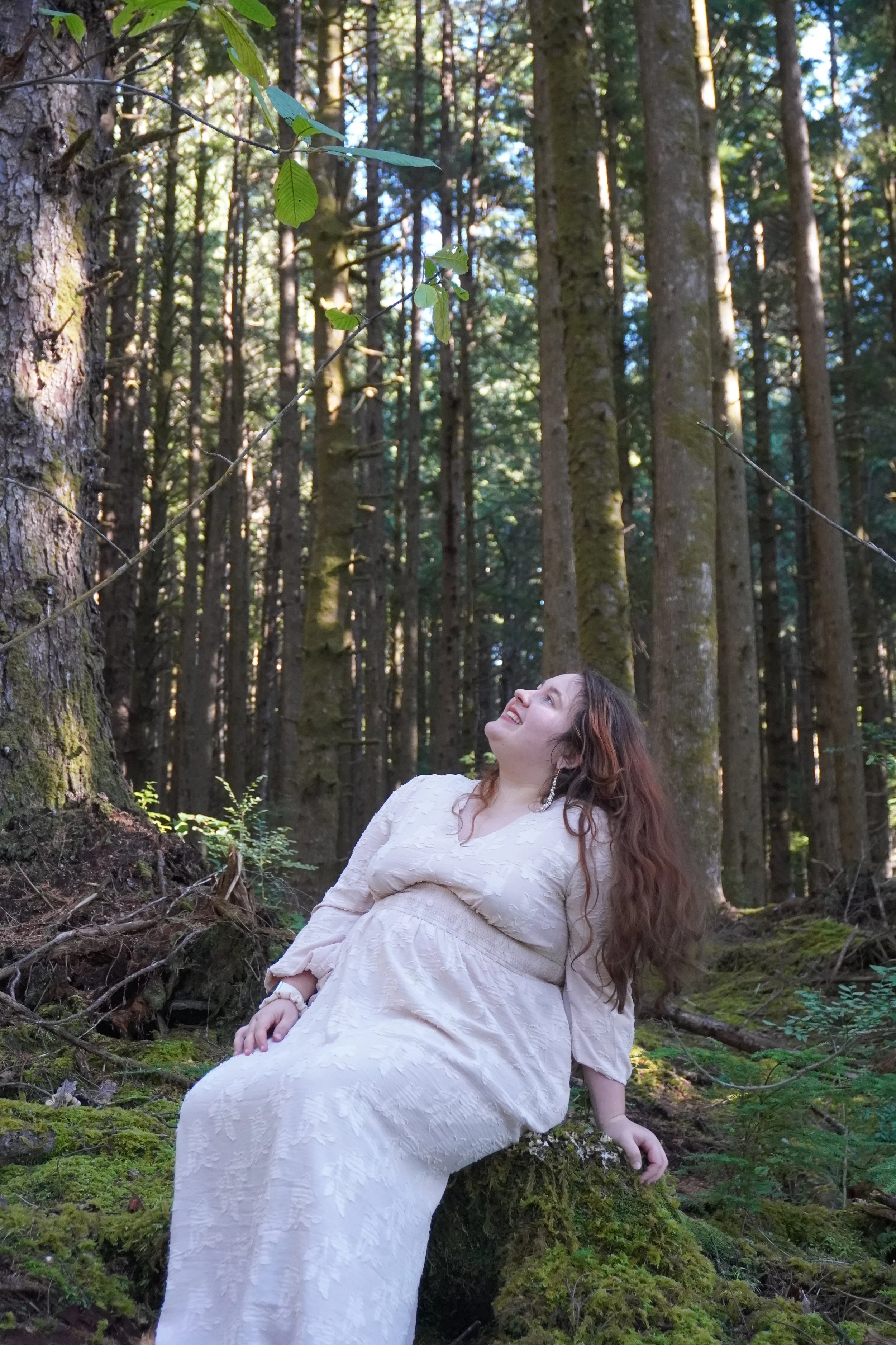 A woman in a white dress sitting on a mossy log in a dense forest, looking up and smiling.