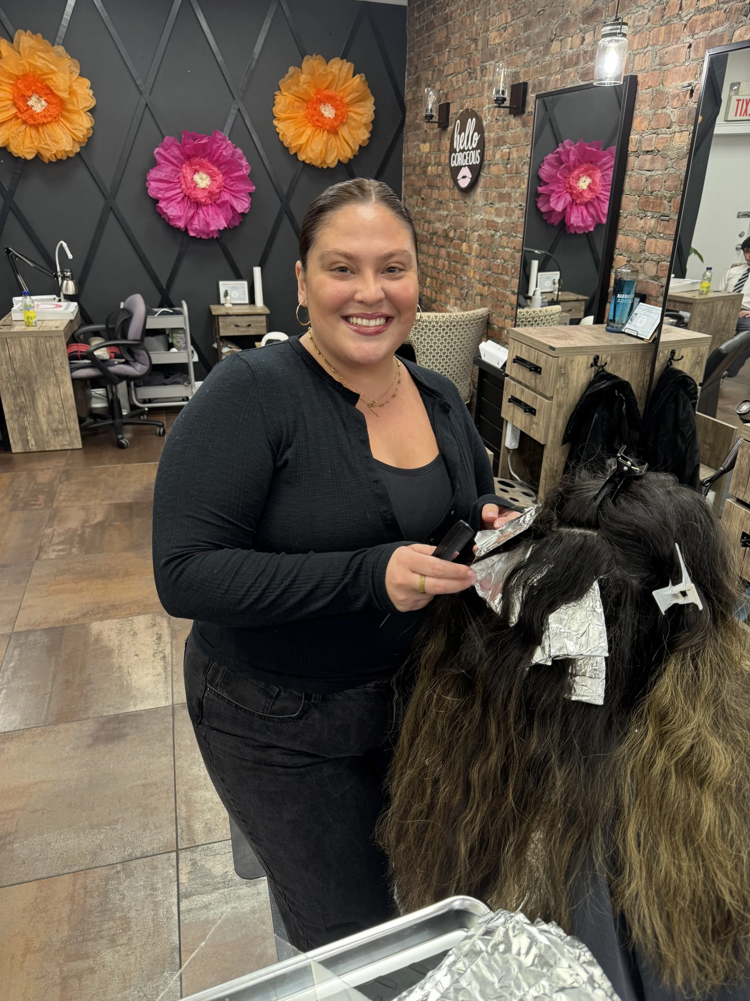A hairstylist with curly blonde hair, wearing a black dress, is applying hair color to a woman's hair using a brush. The woman has shoulder-length brown hair and is seated in a salon chair wearing protective glasses and a black cape. There is a bowl of hair dye on a tray nearby.