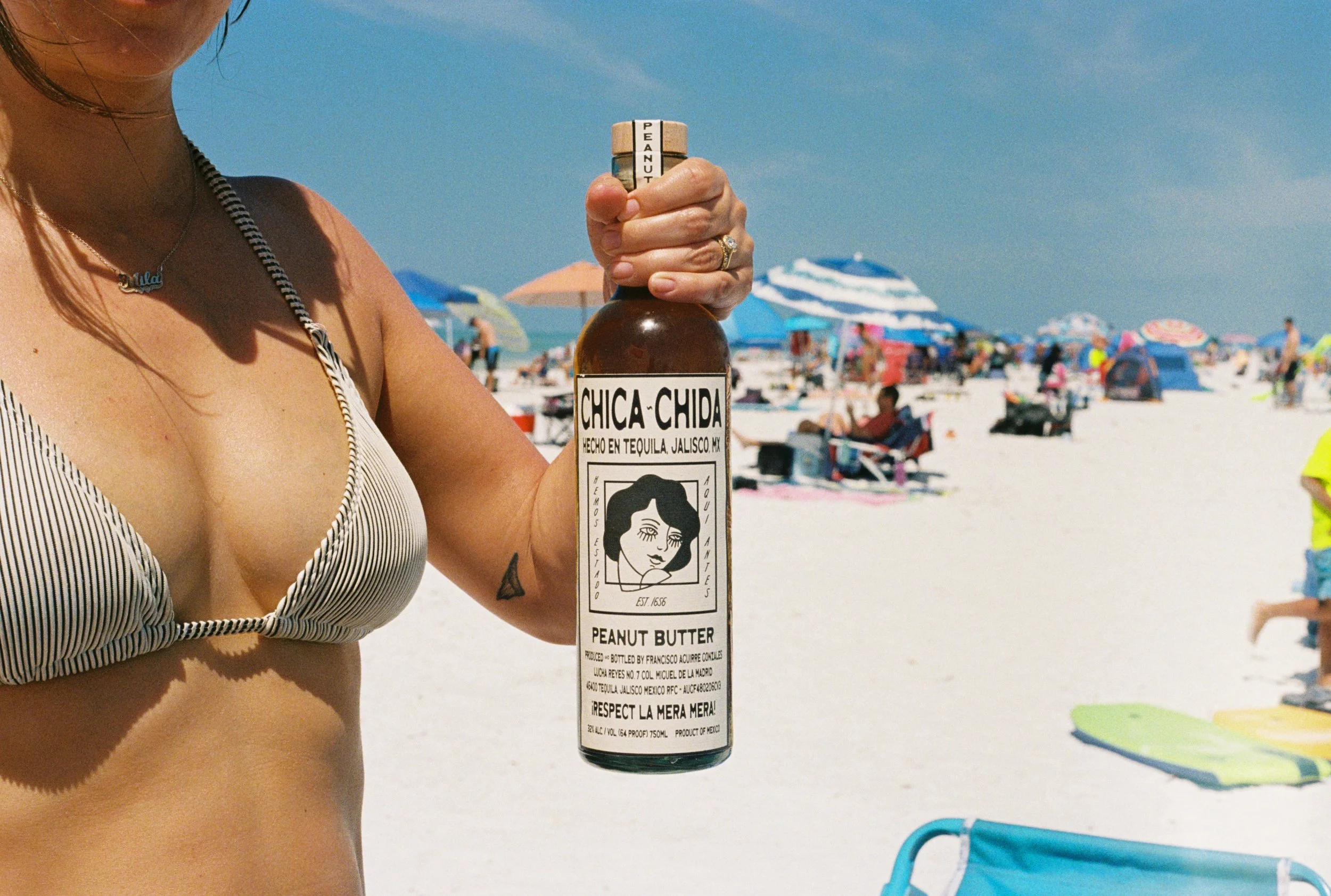 A woman in a striped bikini top holding a bottle of Chica Chida peanut butter tequila at the beach, with umbrellas and people in the background.