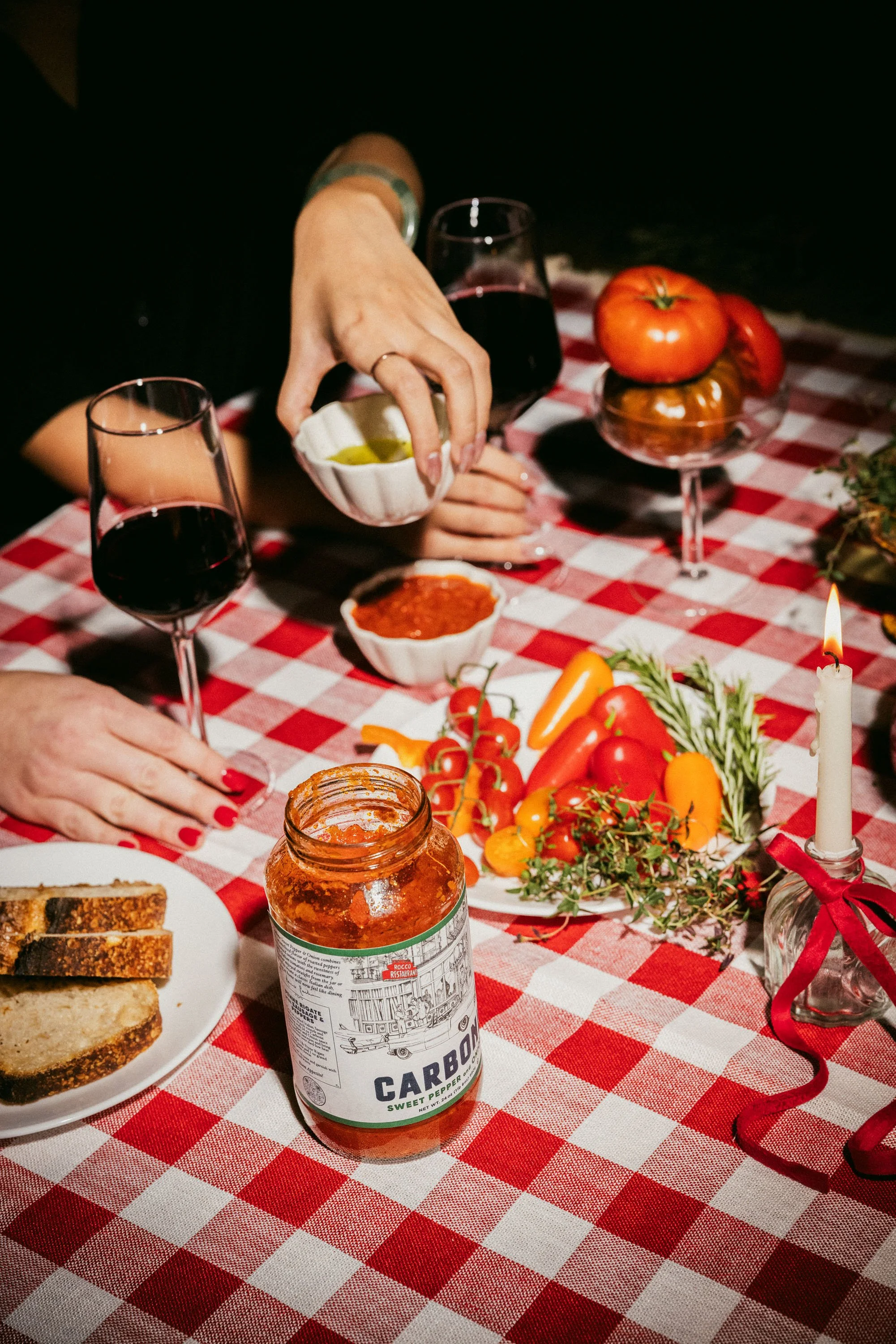 Table with red and white checkered tablecloth, food, wine glasses, tomatoes, bread, jar of pepper, and lit candle