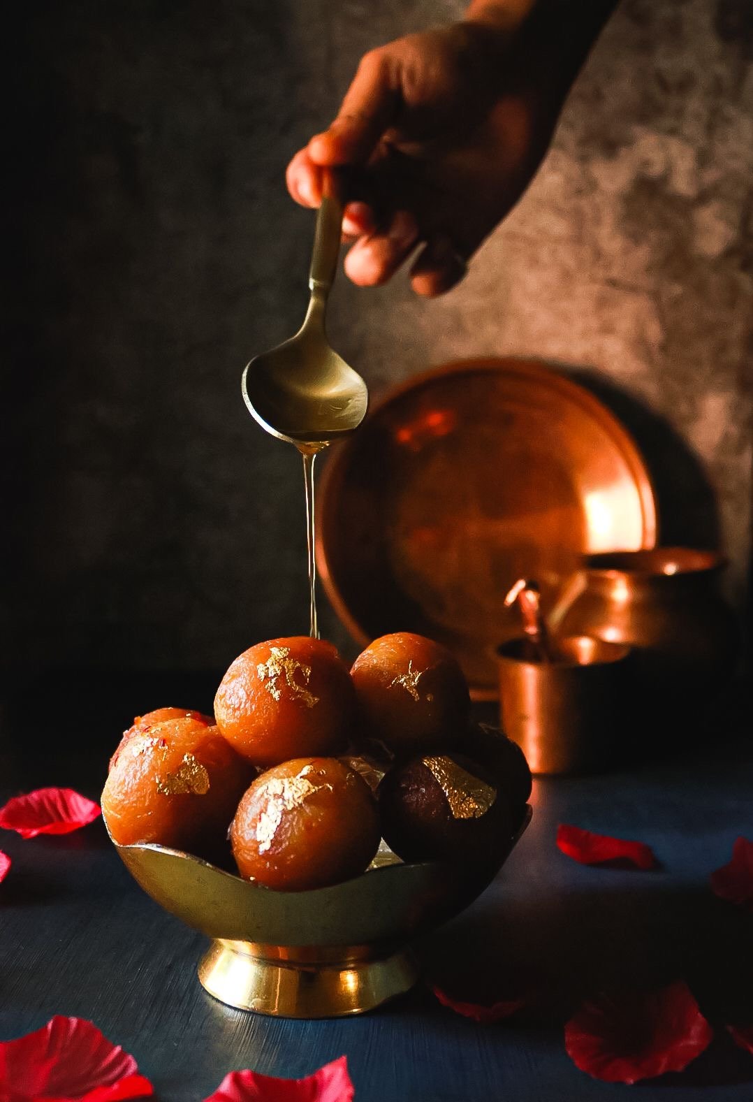 A hand drizzling honey over Indian sweets called gulab jamuns in a golden bowl, with red flower petals scattered around on a dark surface.