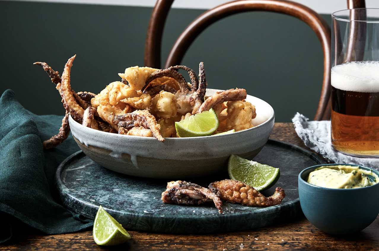A bowl of fried calamari with lemon wedges on a dark serving tray, a glass of beer, and a bowl of dipping sauce on a wooden table with a green cloth napkin.