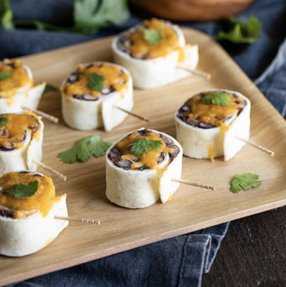 Six mini black bean and cheese roll-ups garnished with cilantro on a wooden serving board.