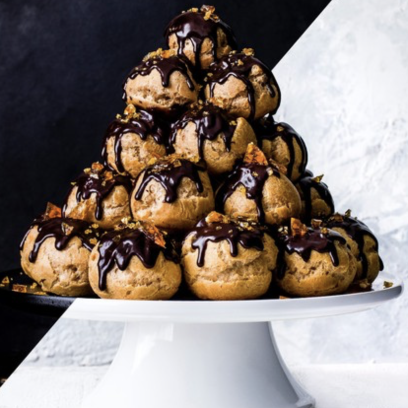 A pyramid of cream puffs topped with chocolate glaze and garnished with small orange-colored bits, displayed on a white cake stand.