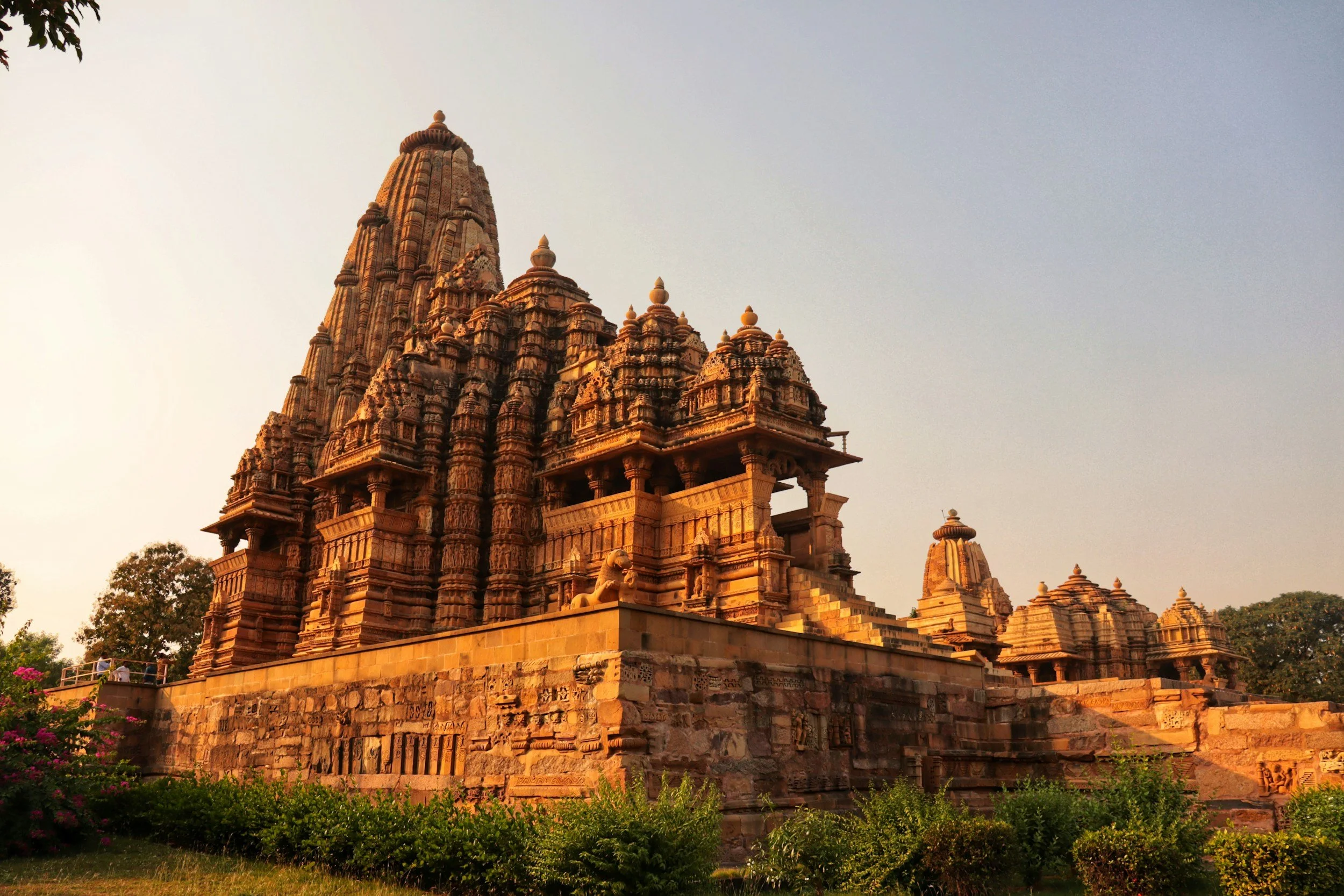 View of a large ancient Hindu temple made of red sandstone, with intricate carvings, multiple spires, and a lion statue at the entrance, set against a clear sky.