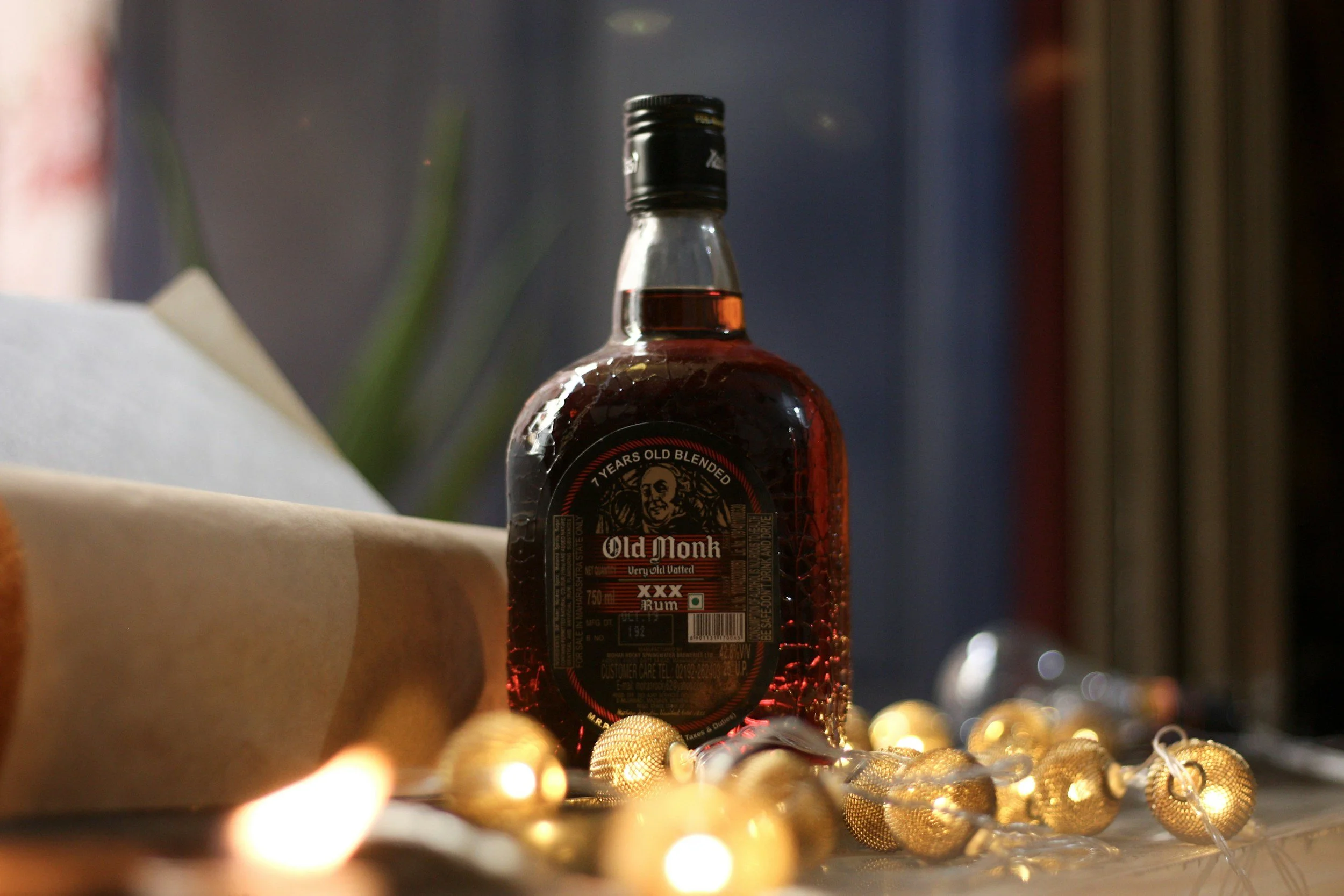 A bottle of Old Monk rum surrounded by gold Christmas ornaments and string lights, with a blurred plant in the background.