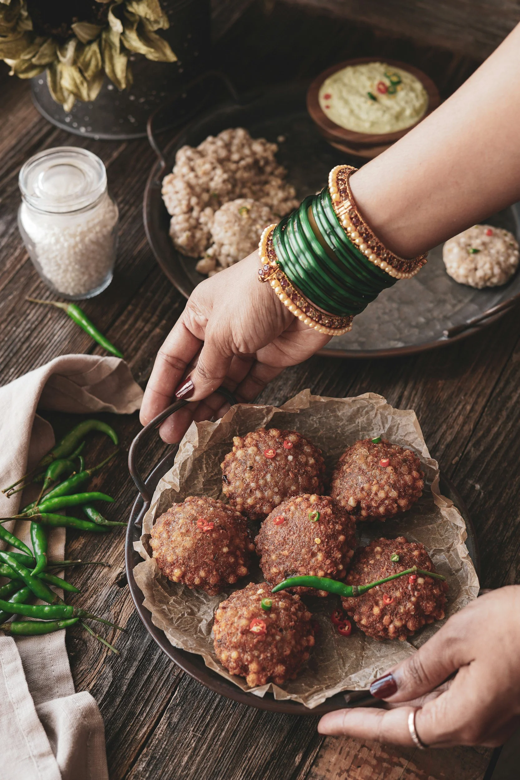 Plate of Indian-style falafel balls garnished with sliced red chili, on a rustic wooden table with green chili peppers and a jar of white sauce nearby.