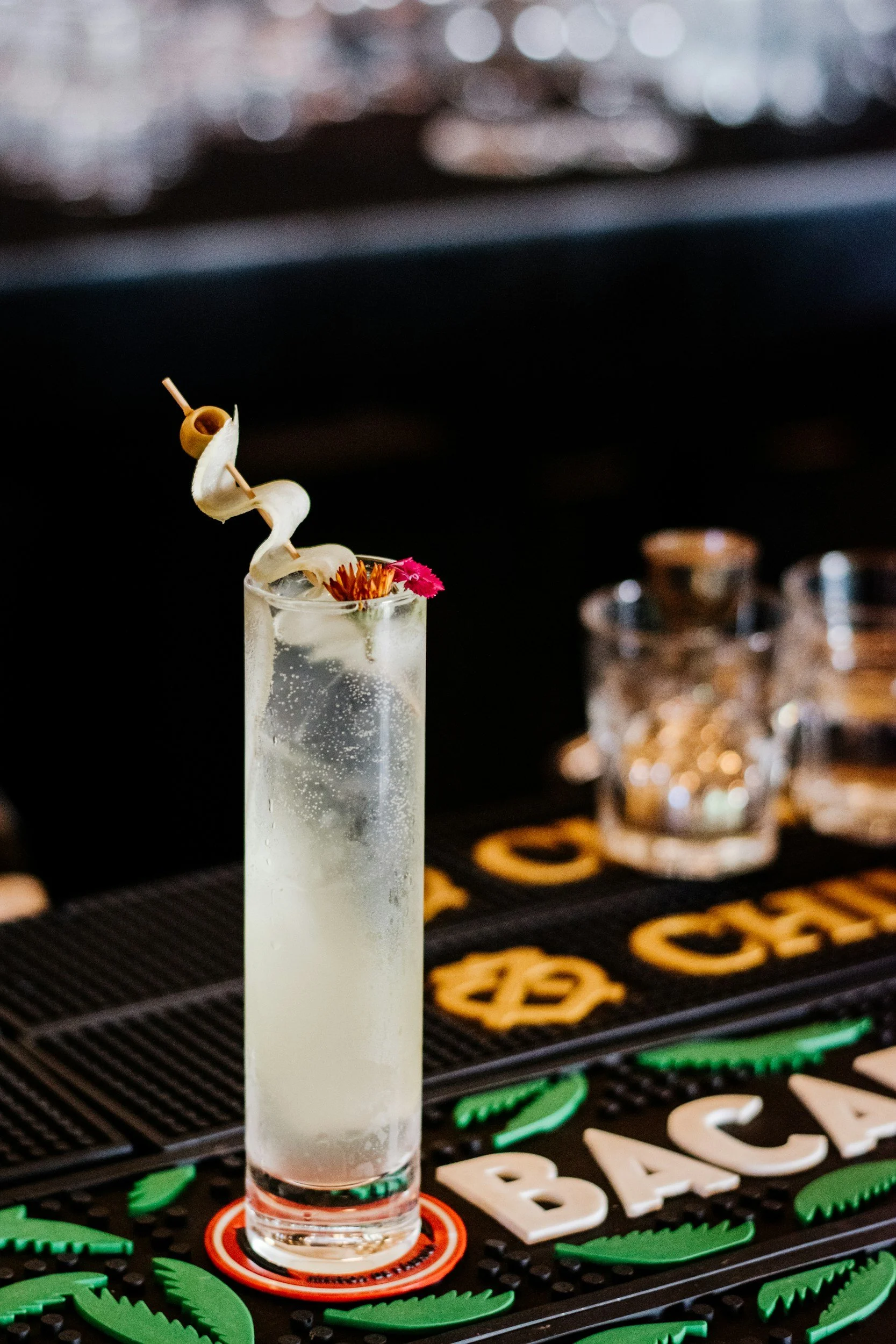 A tall glass of cocktail with a twist of banana peel and garnished with a pink flower on a bar counter.