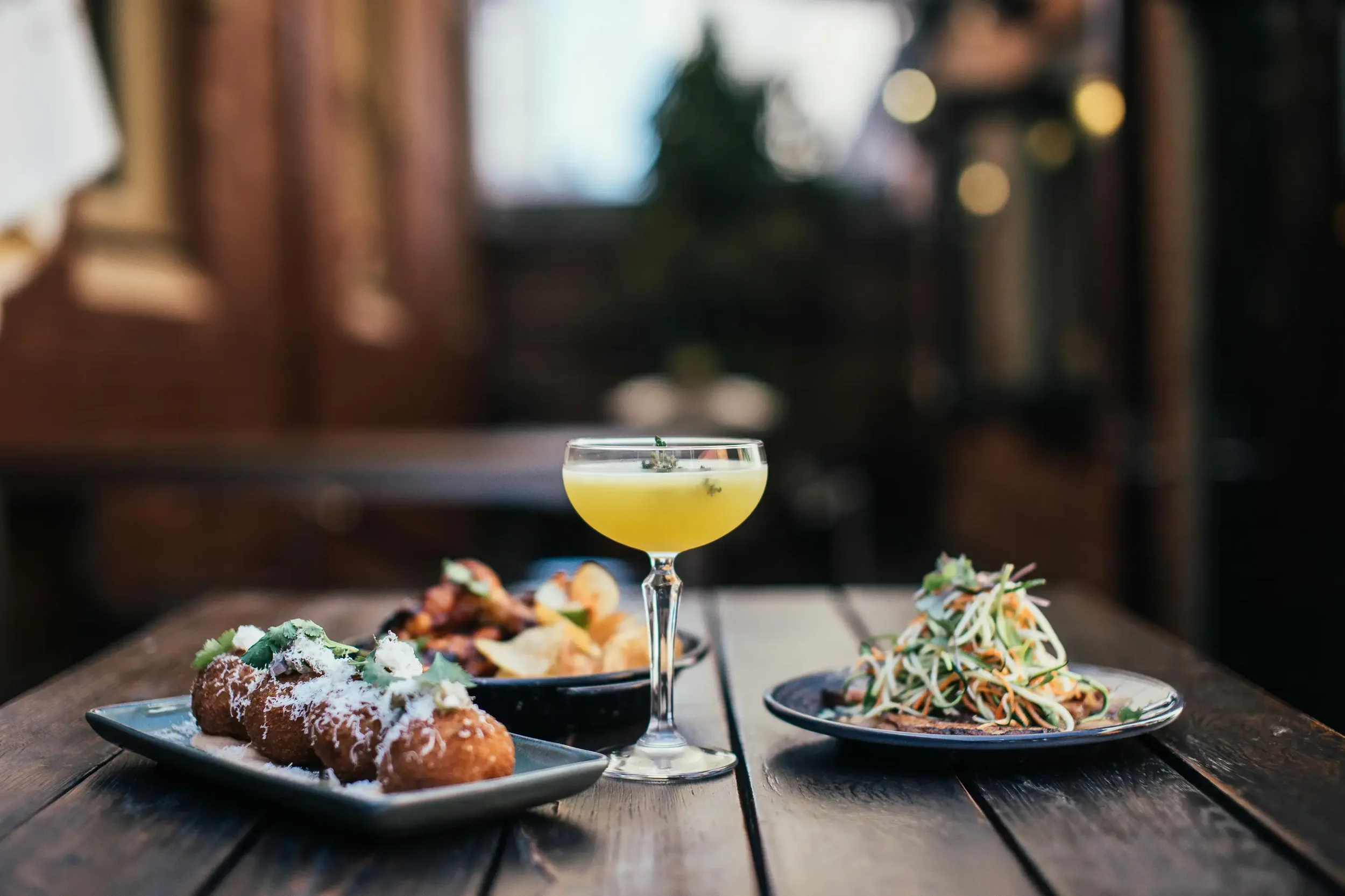 Appetizers and a cocktail on a wooden table in a dimly lit restaurant.