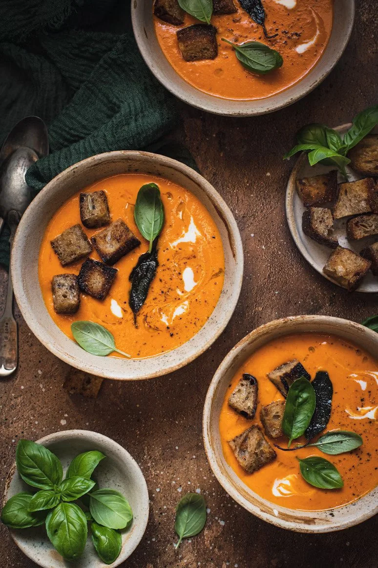 Bowl of creamy tomato or butternut squash soup topped with grilled cubes, basil leaves, black pepper, and a swirl of cream. Additional bowls with similar soup and toppings are visible, along with small bowls of fresh basil leaves.