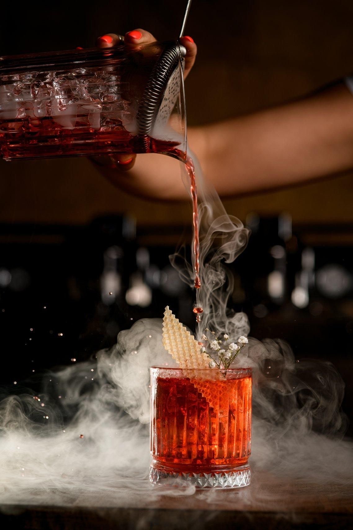 A hand pouring red liquid into a glass with decorative elements, surrounded by atmosphere with fog or steam.