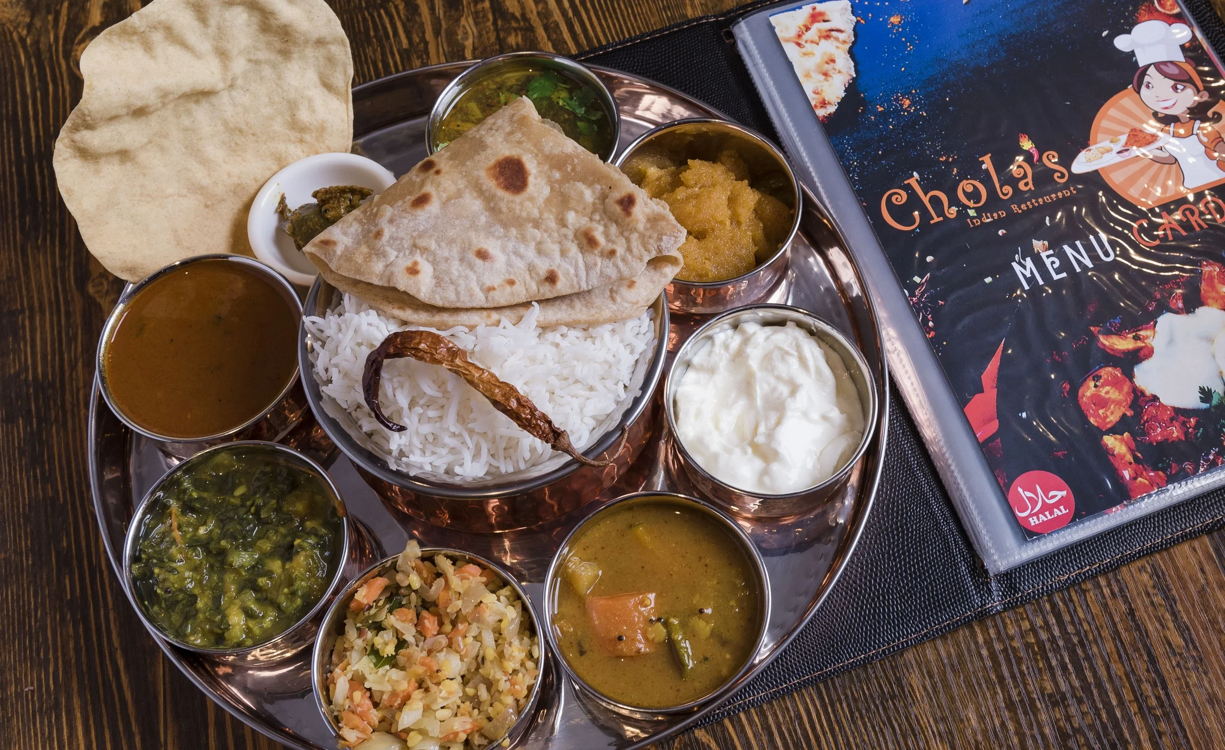 Traditional Indian thali with rice, naan bread, various curries and side dishes, and a glass of yogurt, served on a metal tray.