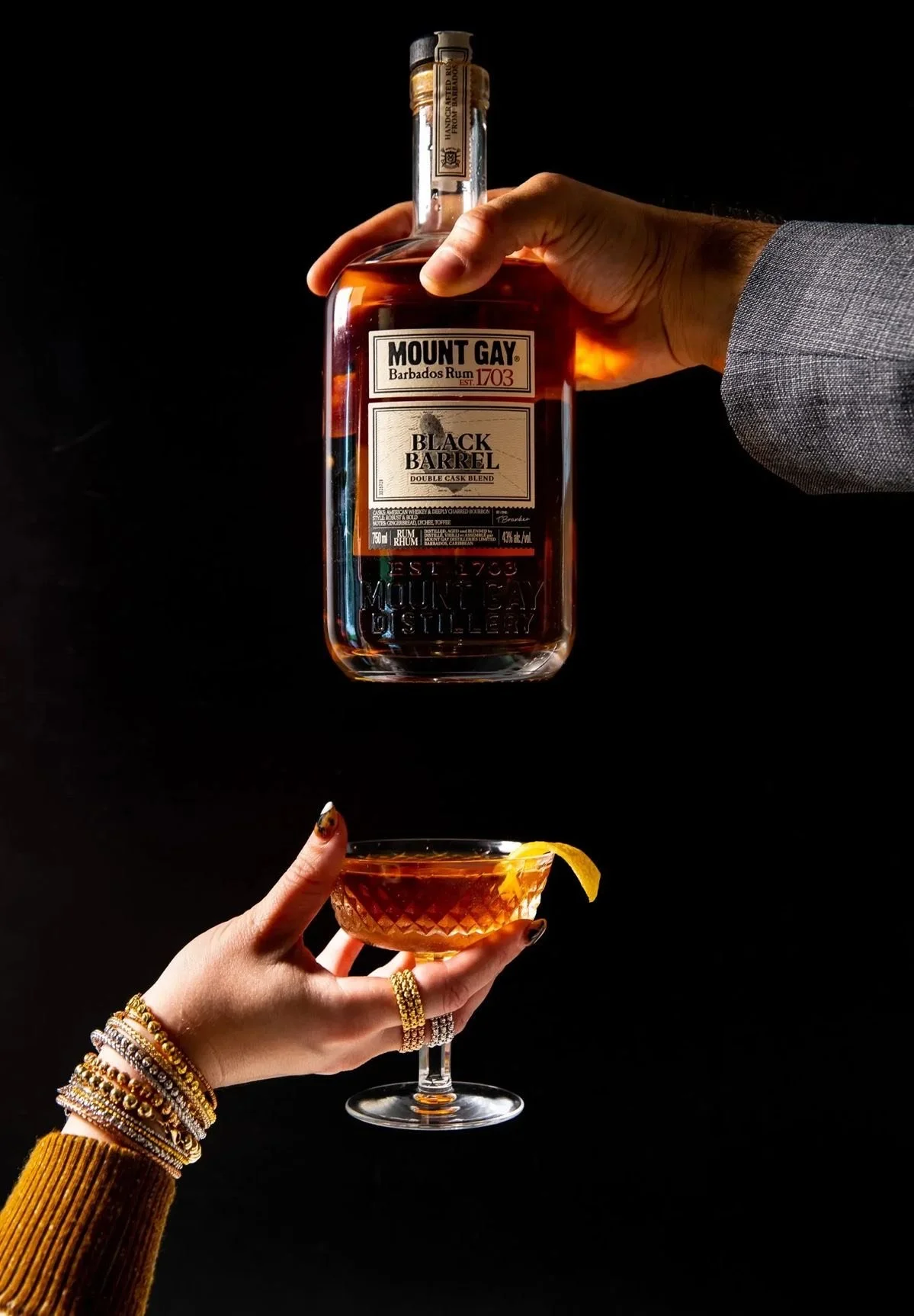 Hand holding a bottle of Mount Gay Black Barrel rum above a glass with a cocktail garnished with a lemon twist against a black background.