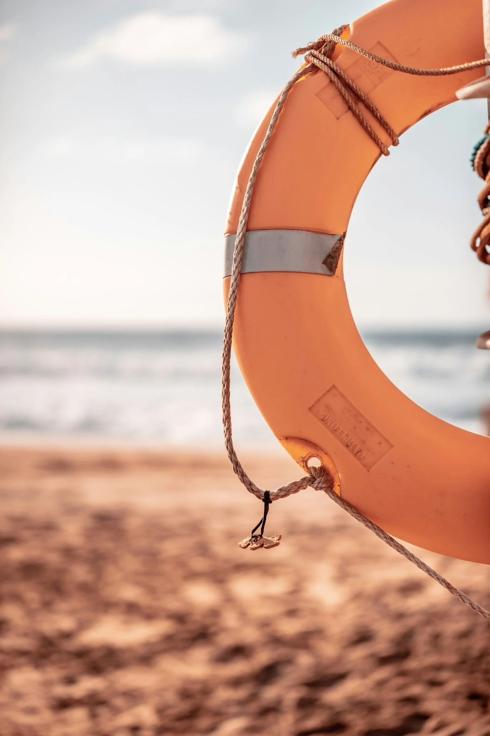 Orange life preserver hanging on a rope on a sandy beach with ocean waves in the background, under cloudy sky.