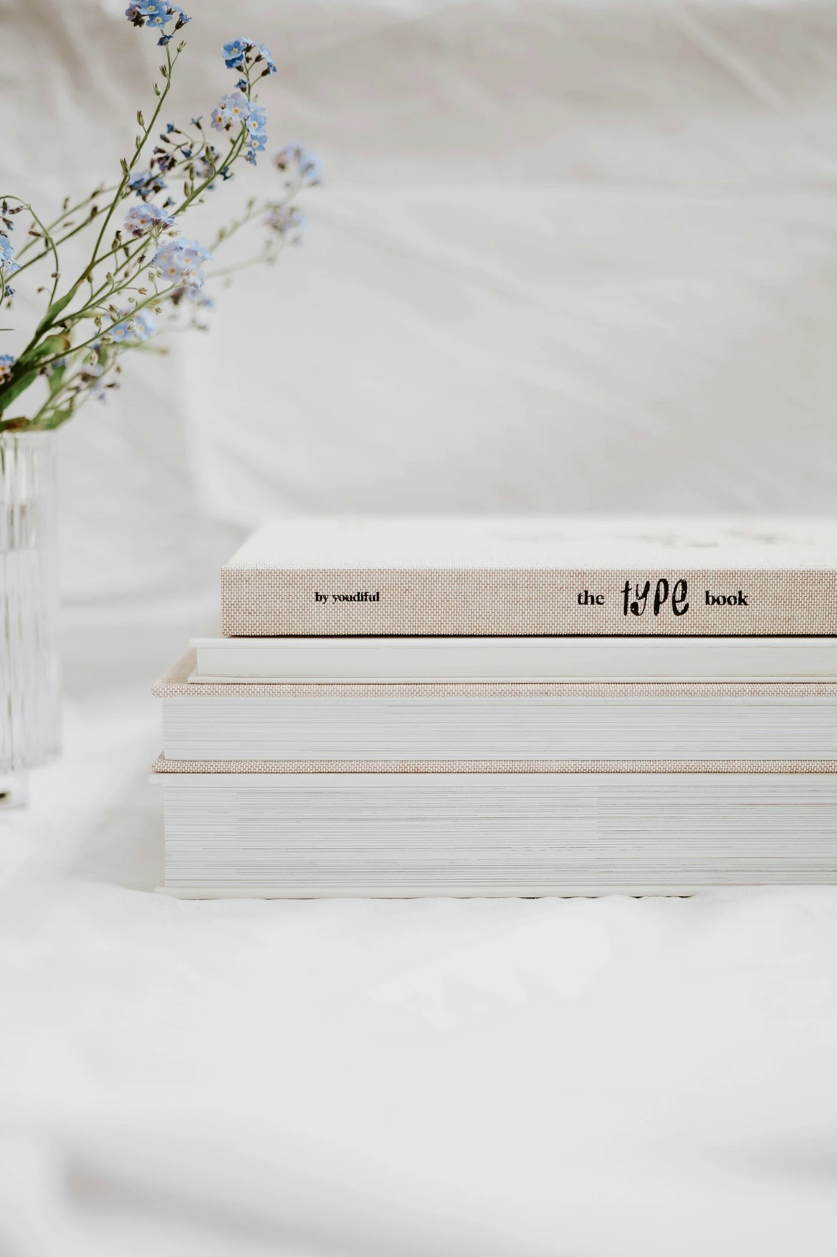 A stack of four books with minimalist covers sits on a white surface, with a vase of small blue and white flowers on the left side.