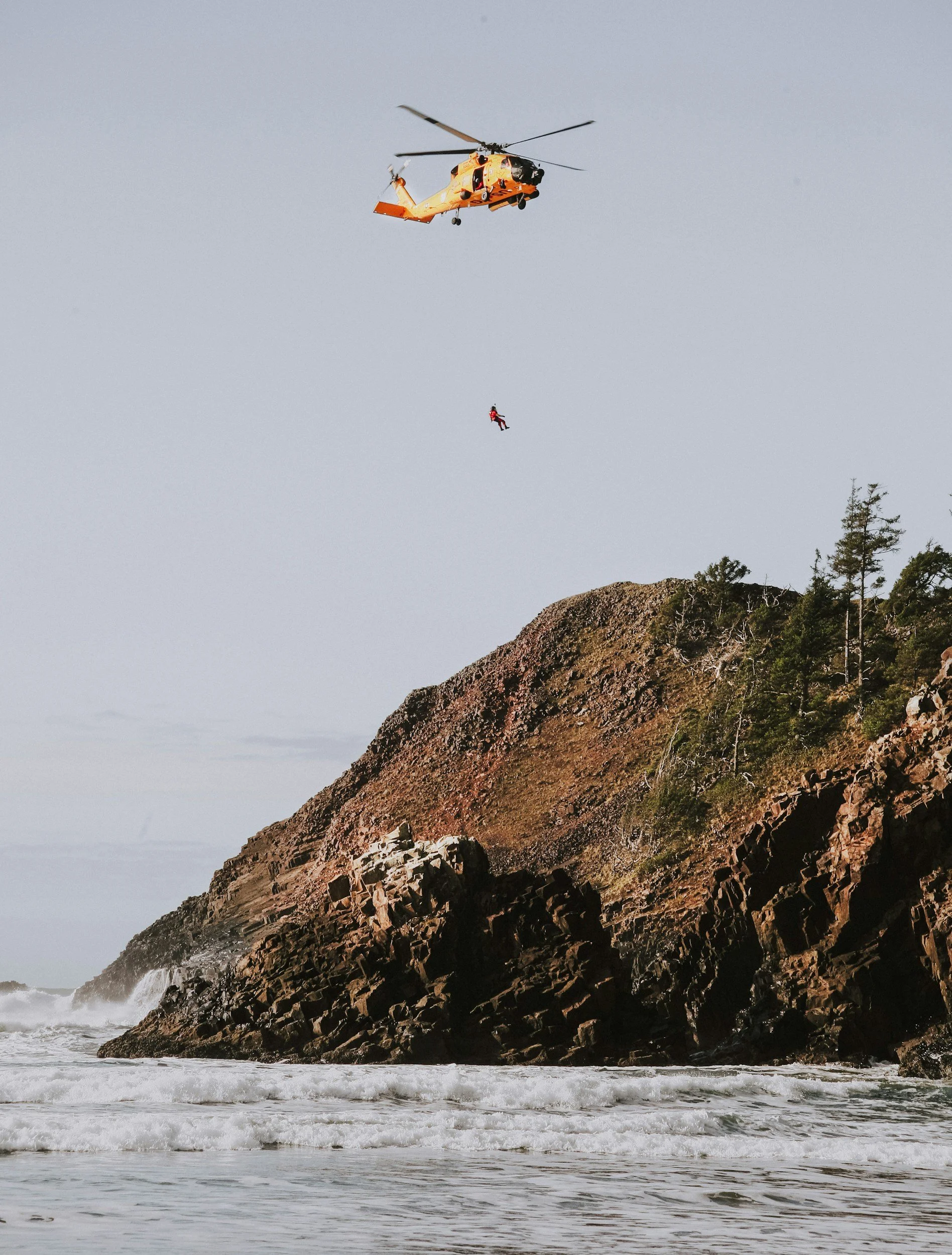 A helicopter hovering above the ocean coastline with a person being hoisted up in a rescue harness, near a rocky cliff with trees.