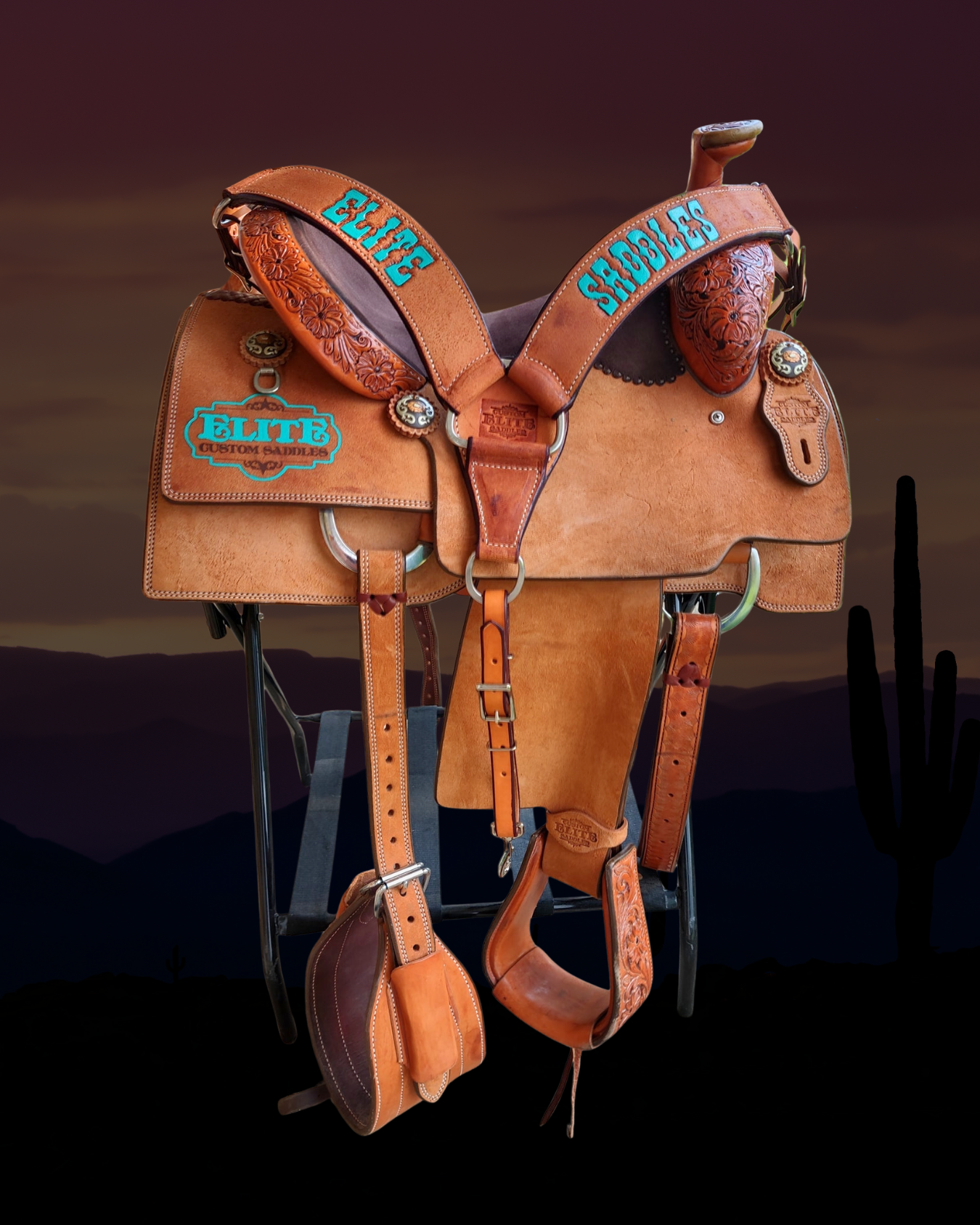 A brown Western saddle with turquoise embroidered details is mounted on a saddle stand against a desert sunset background with silhouettes of cacti and mountains.