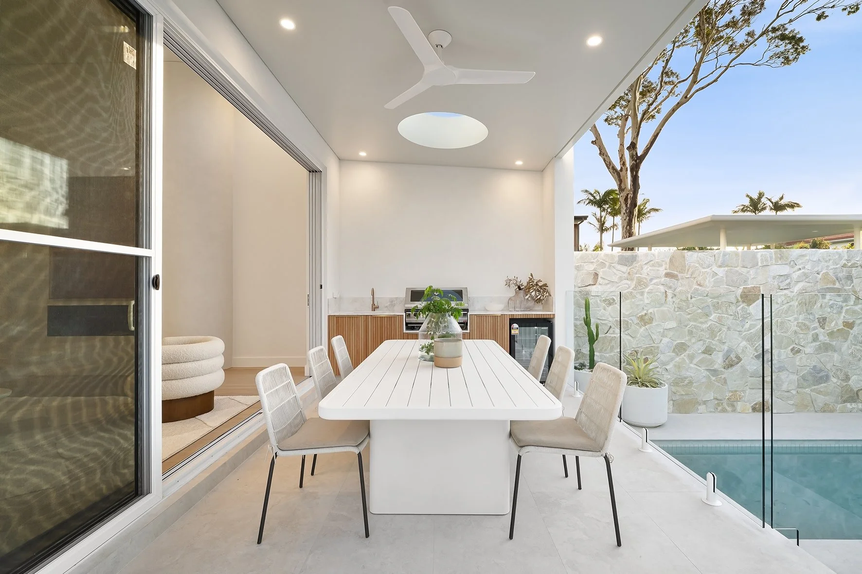 Modern outdoor dining area with a white rectangular table, six beige chairs, and a decorated table with a vase of greenery. The area is enclosed by a glass windbreak, with a stone wall, potted plants, trees, and a swimming pool visible beyond.