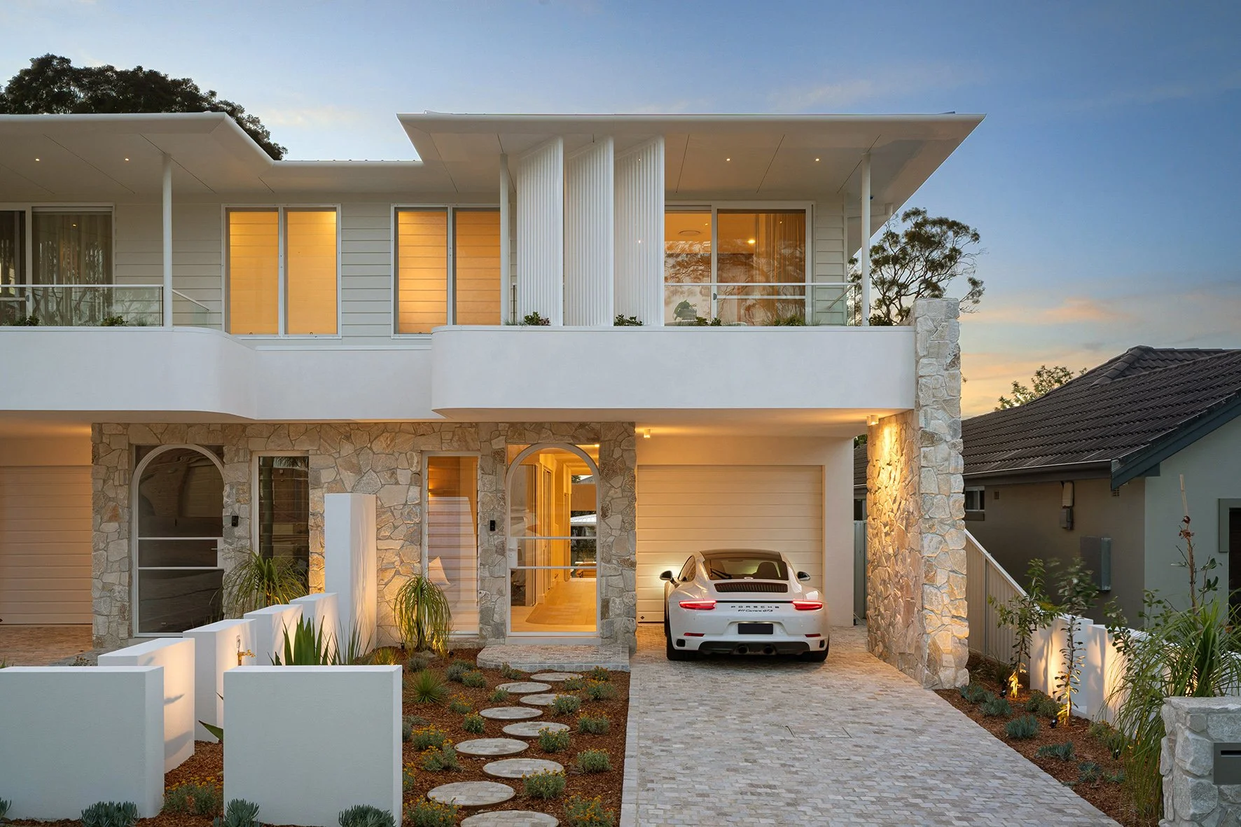 Modern multi-story house with stone and white exterior, large windows, a car parked in the driveway, and landscaped front yard during dusk.
