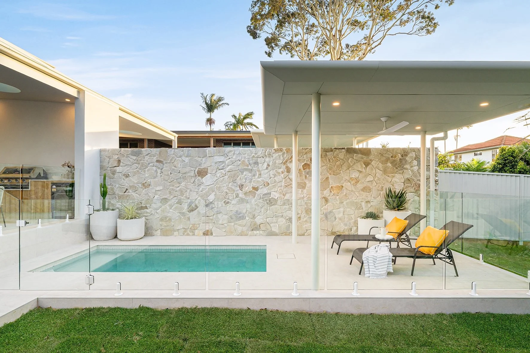 Modern outdoor patio with a pool, lounge chairs with yellow pillows, potted succulents, stone wall, glass fencing, and a shaded area with a ceiling fan and additional chairs, in a residential backyard.