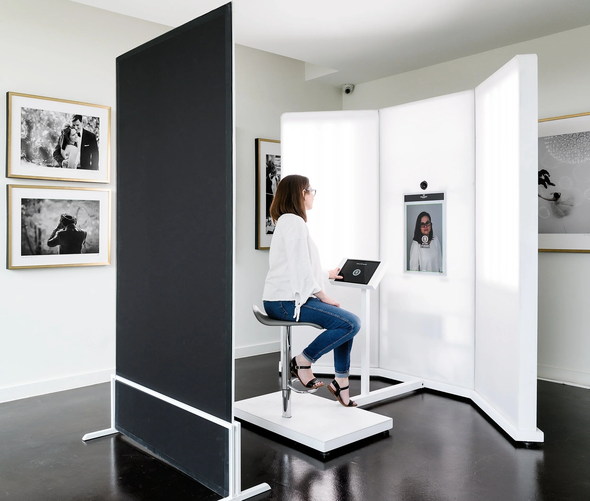A woman sitting on a stool is having her photograph taken by a face-scanning device in an art gallery.
