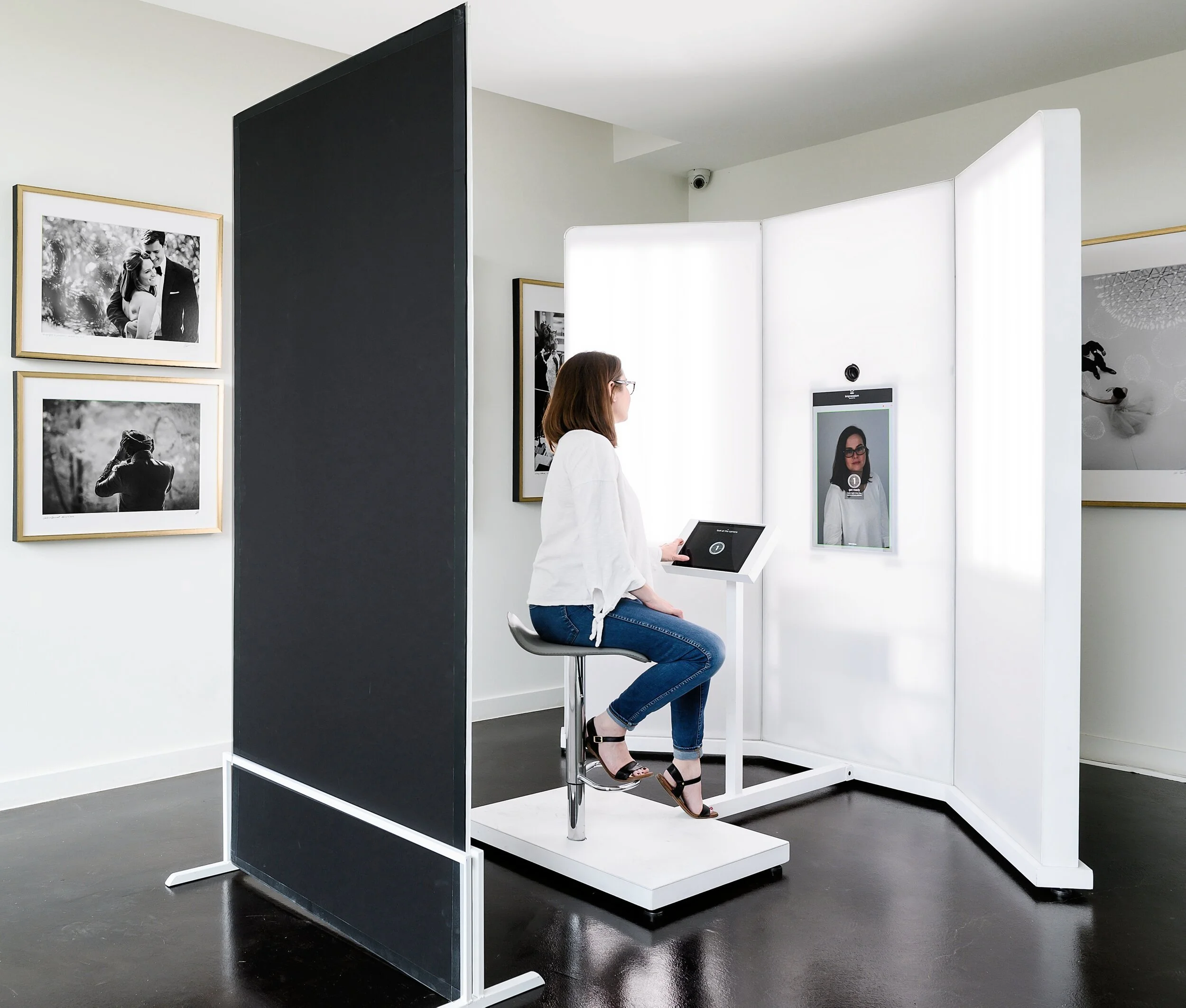 A woman is sitting on a stool in a photo booth with a digital screen displaying her portrait, inside an art gallery. The photo booth has white, bright lighting panels, and black and white framed photographs are on the gallery walls.