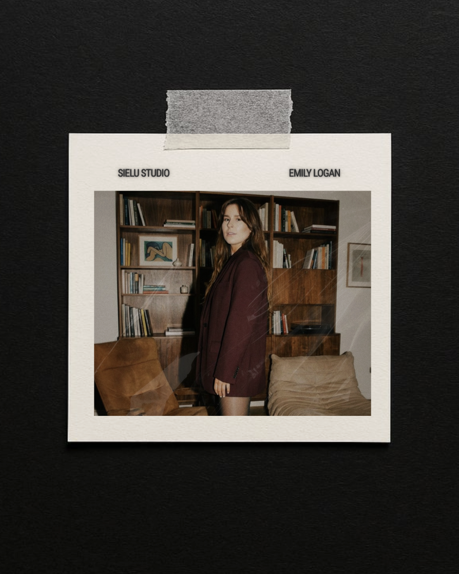 Photograph of a young woman in a burgundy blazer standing in a room with wooden bookshelves and beige furniture, framed and taped to a black background.