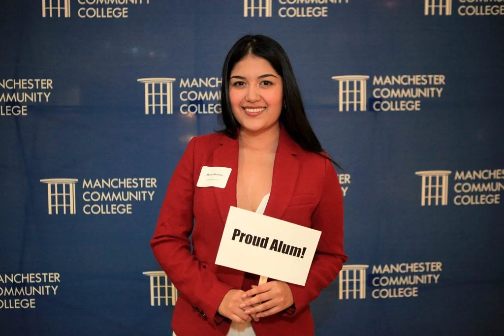 Woman holding a sign that reads "Proud Alum" in front of a Manchester Community College backdrop