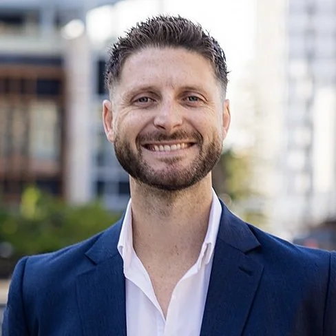 Smiling man in blue suit and white shirt, standing outdoors with blurred buildings in the background.