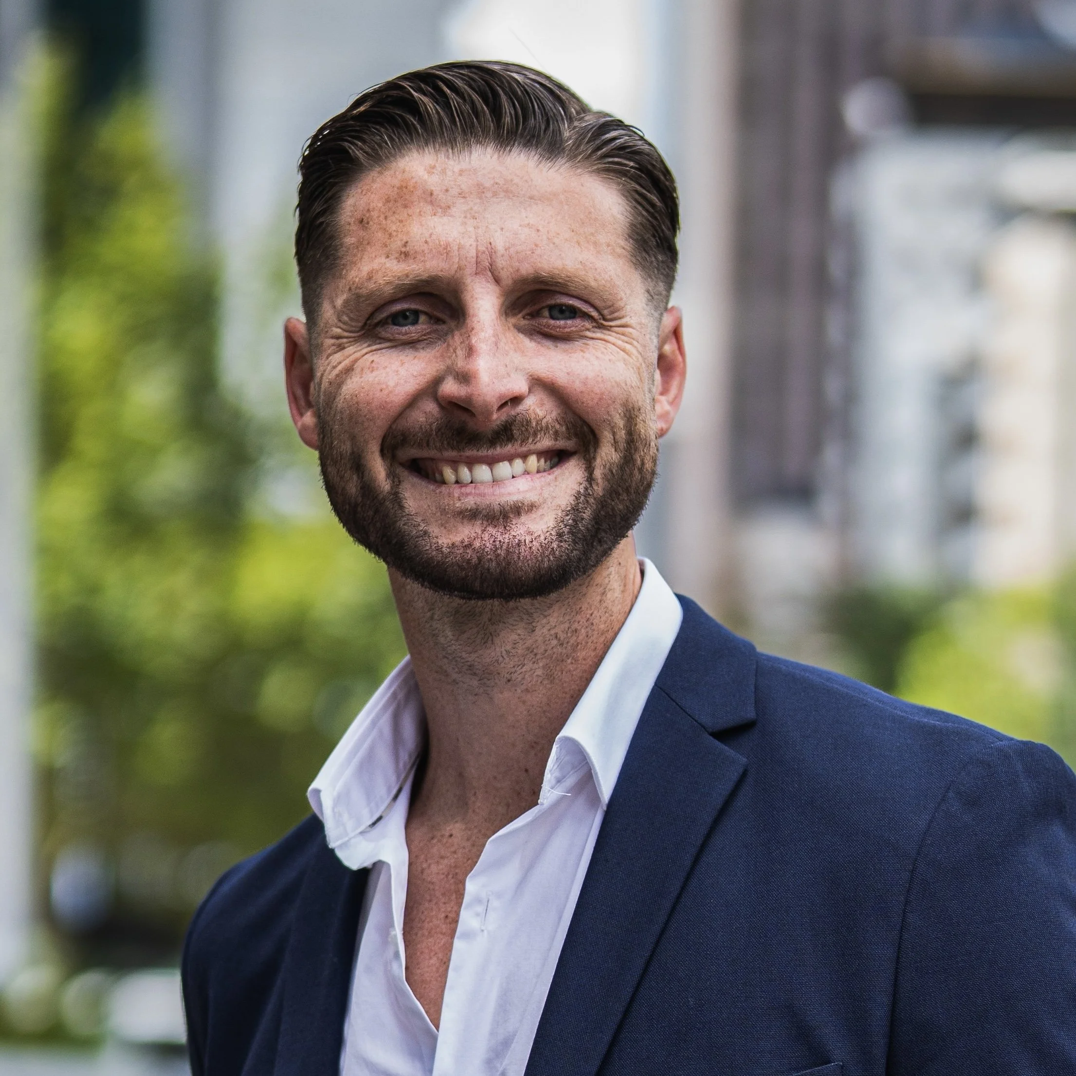 Smiling man in a blue suit standing outdoors with blurry buildings in the background.