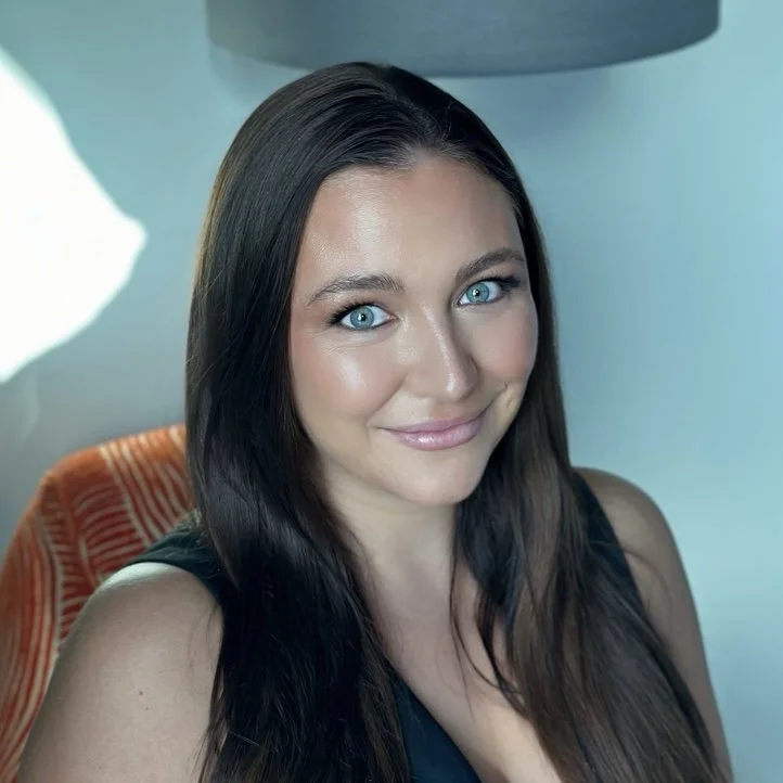 A woman with long brown hair smiling, wearing a black top, sitting indoors with an upholstered chair and a lampshade in the background.