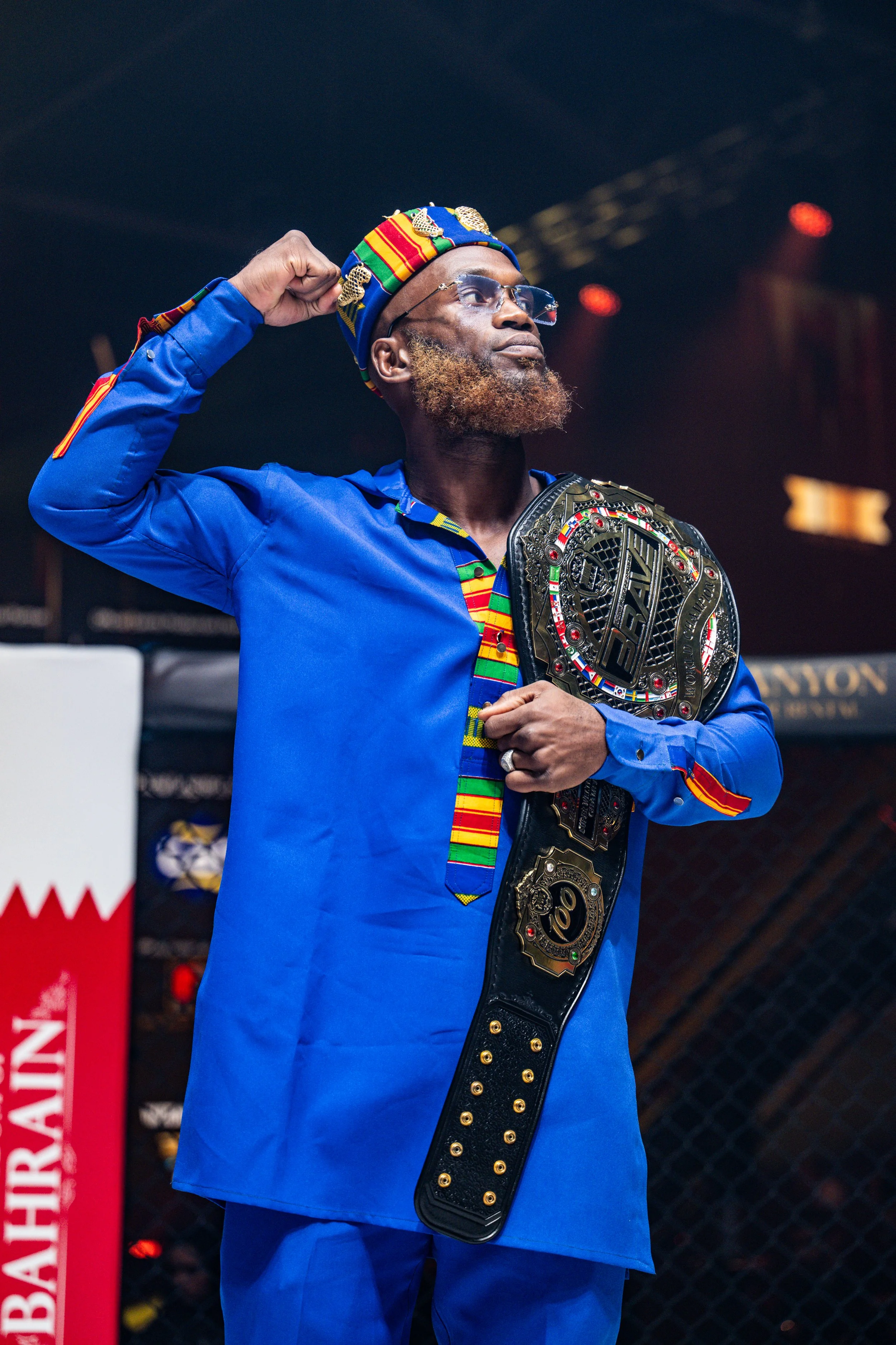 A man in a blue outfit with colorful patterns, wearing glasses and a colorful hat, holding a championship belt over his shoulder in an arena setting.