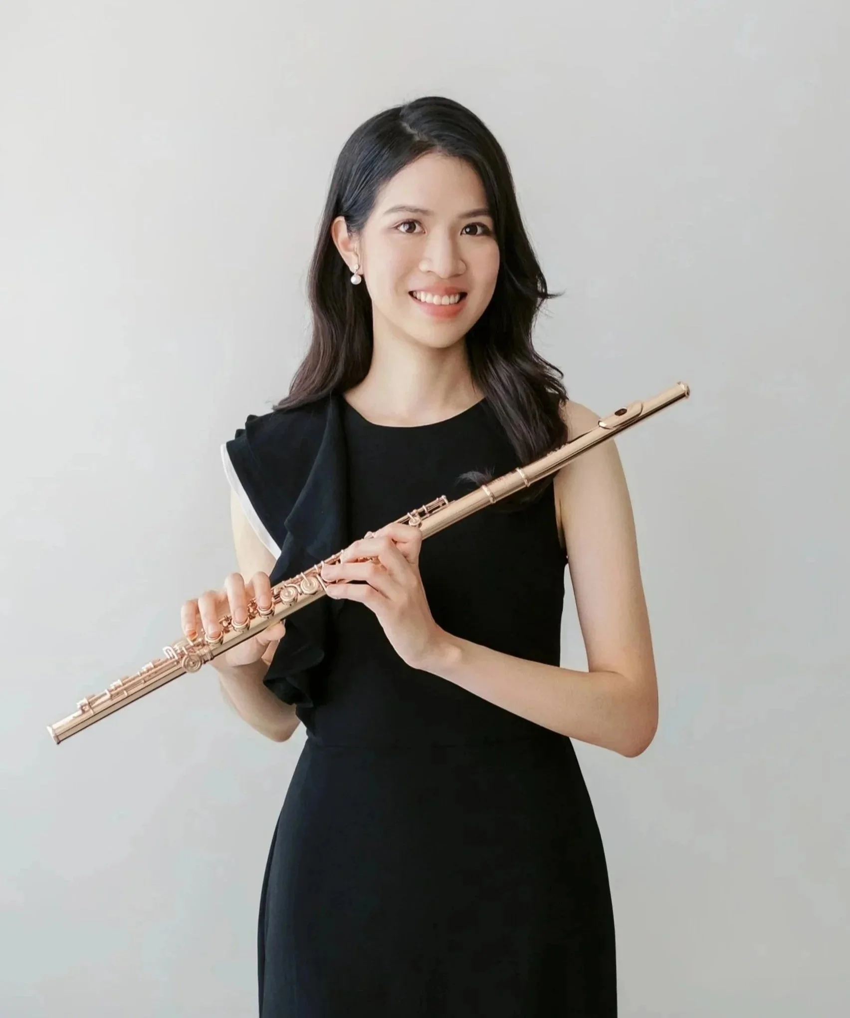 A young woman with dark hair wearing pearl earrings and a black dress, holding a flute, smiling, standing against a plain white background.