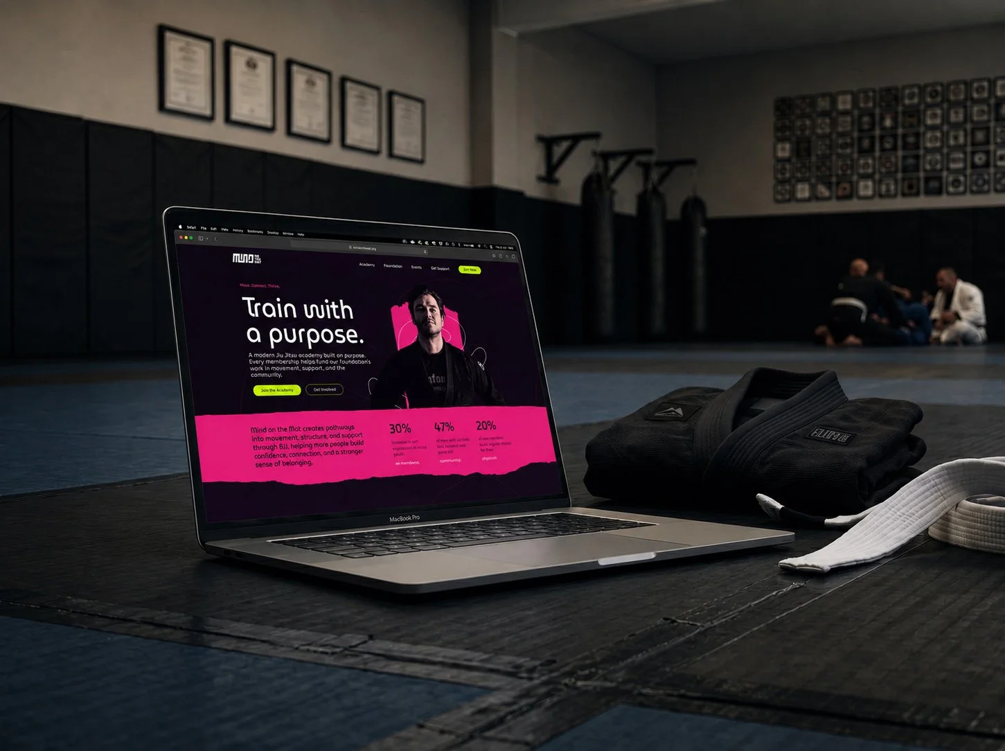 A laptop on the floor in a martial arts gym displaying a website with the slogan "Train with a purpose". Nearby are martial arts gloves and belt, with three people practicing Brazilian Jiu-Jitsu in the background.