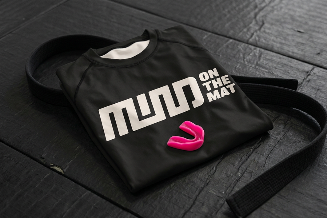 A black martial arts uniform with white text that reads 'MUDO ON THE MAT' lying on a dark wooden surface, with a pink mouth guard placed on top of it, and a black belt partially visible.