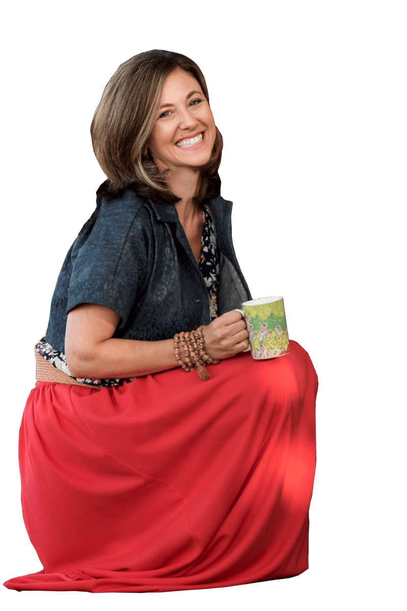 A smiling woman with shoulder-length hair, wearing a dark short-sleeved top, a long red skirt, multiple bracelets, and holding a floral coffee mug. She is sitting cross-legged and facing slightly to the right.