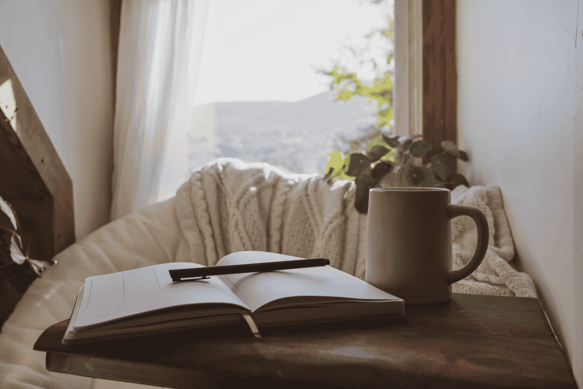 Open book with a black pen on top, a white mug, and a cozy blanket on a bed in front of a window with white curtains, sunlight streaming in, and a view of trees outside.
